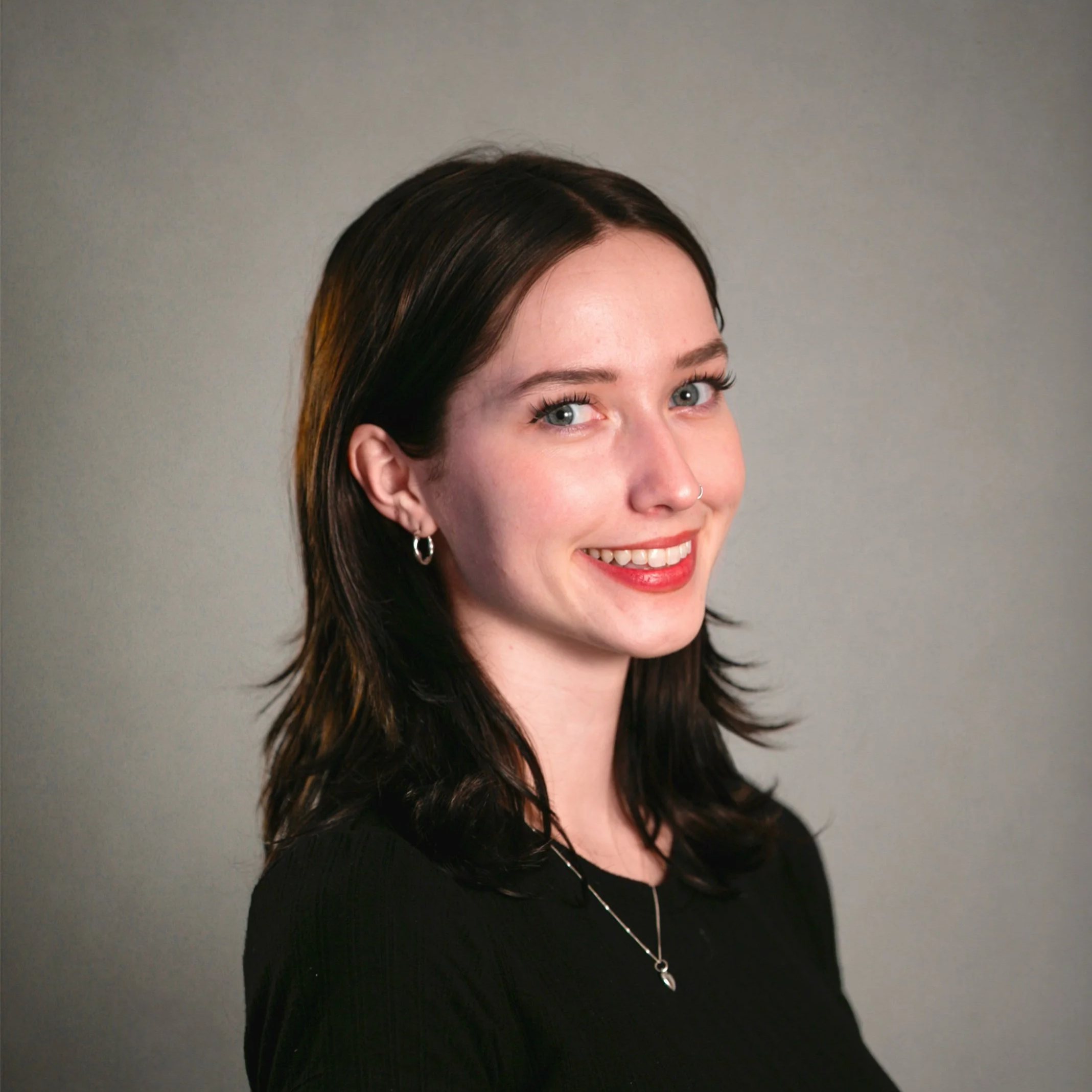 A young woman with dark brown hair, blue eyes, and light skin, smiling and wearing a black top. She has earrings, a nose ring, and a necklace, against a neutral background.