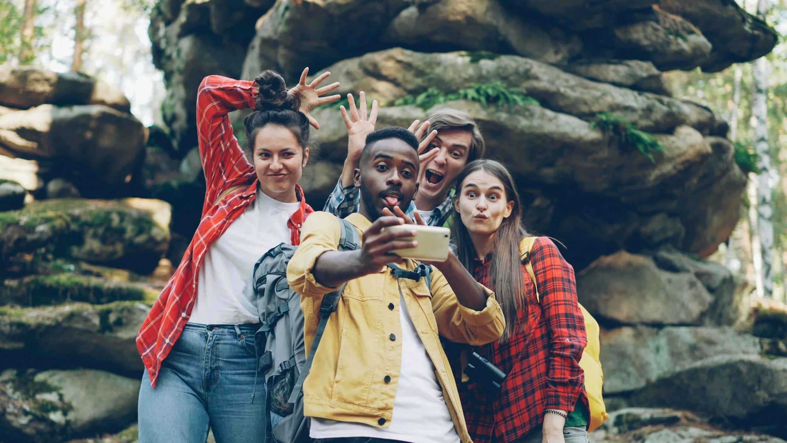 Four friends taking a selfie outdoors in front of large rocks and trees, making funny faces and waving.