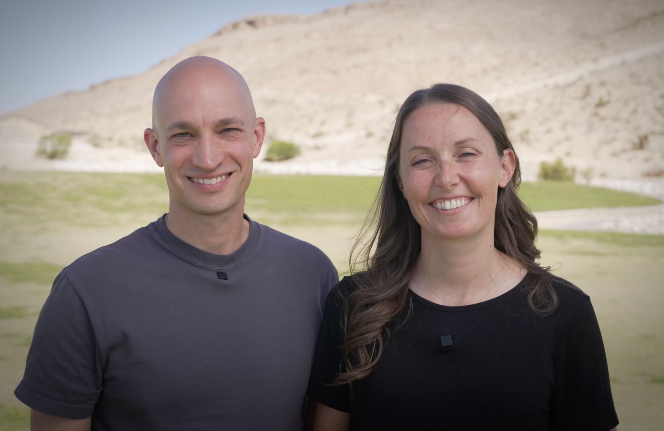 A husband and wife standing outdoors with a rocky hill in the background, smiling.
