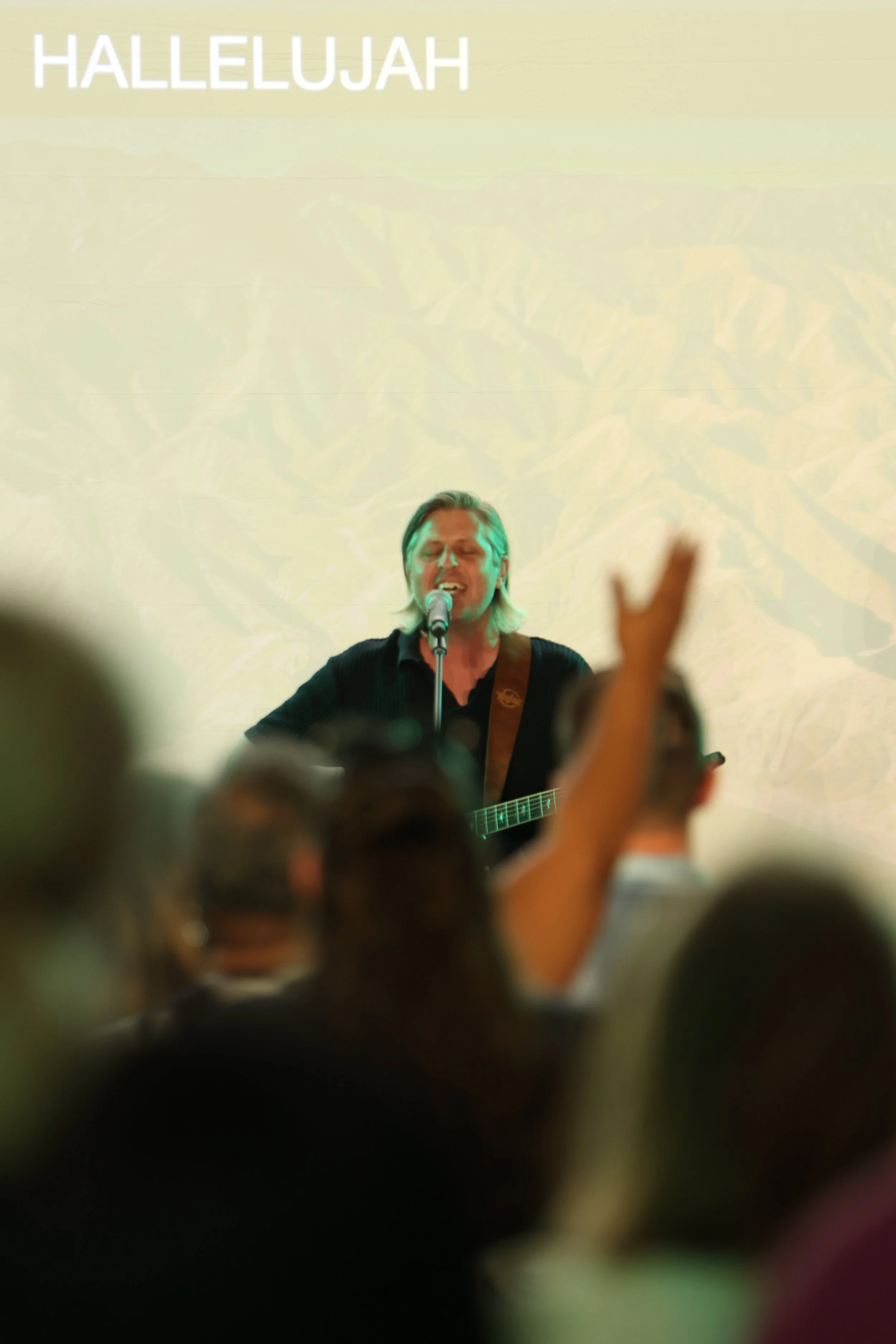 A worship pastor on a stage singing while playing a guitar during a church service. Audience members are visible in the foreground raising their hands in praise and worship.