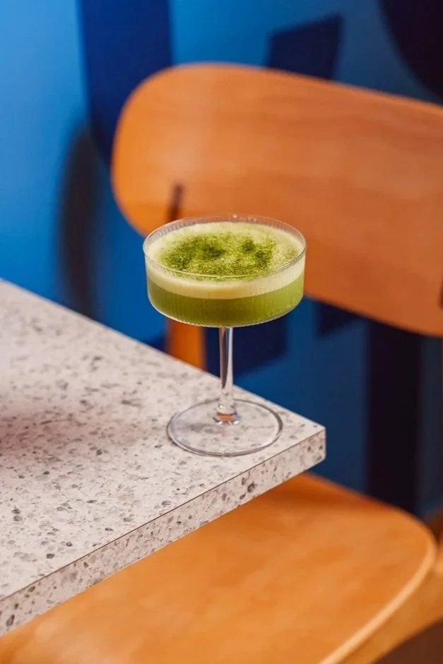 Cóctel verde en copa de cóctel sobre una mesa de granito con silla de madera al fondo.