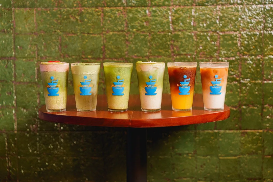 Seis cafés en vasos con diferentes colores y coberturas, sobre una mesa de madera, con fondo de pared verde.