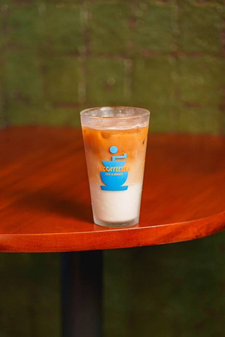Vaso de café con leche y espuma, con un logotipo azul y naranja que dice 'La Coffee Terra' en una mesa de madera.