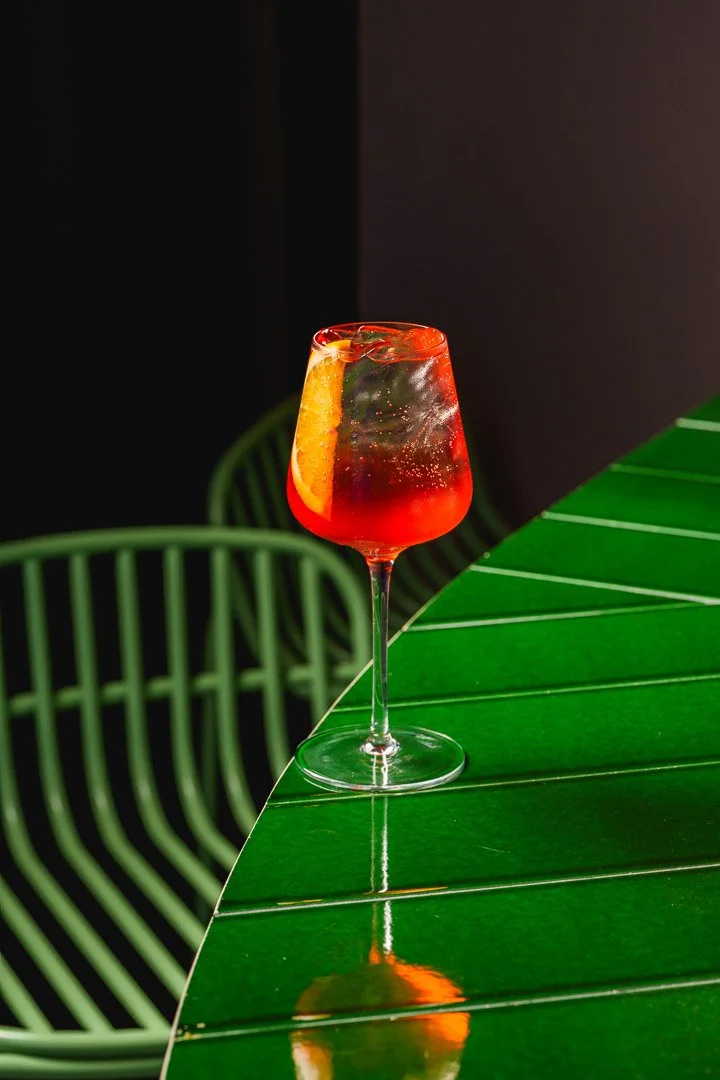 Cóctel rojo con rodaja de naranja en un vaso alto sobre una barra verde con sillas verdes al fondo.