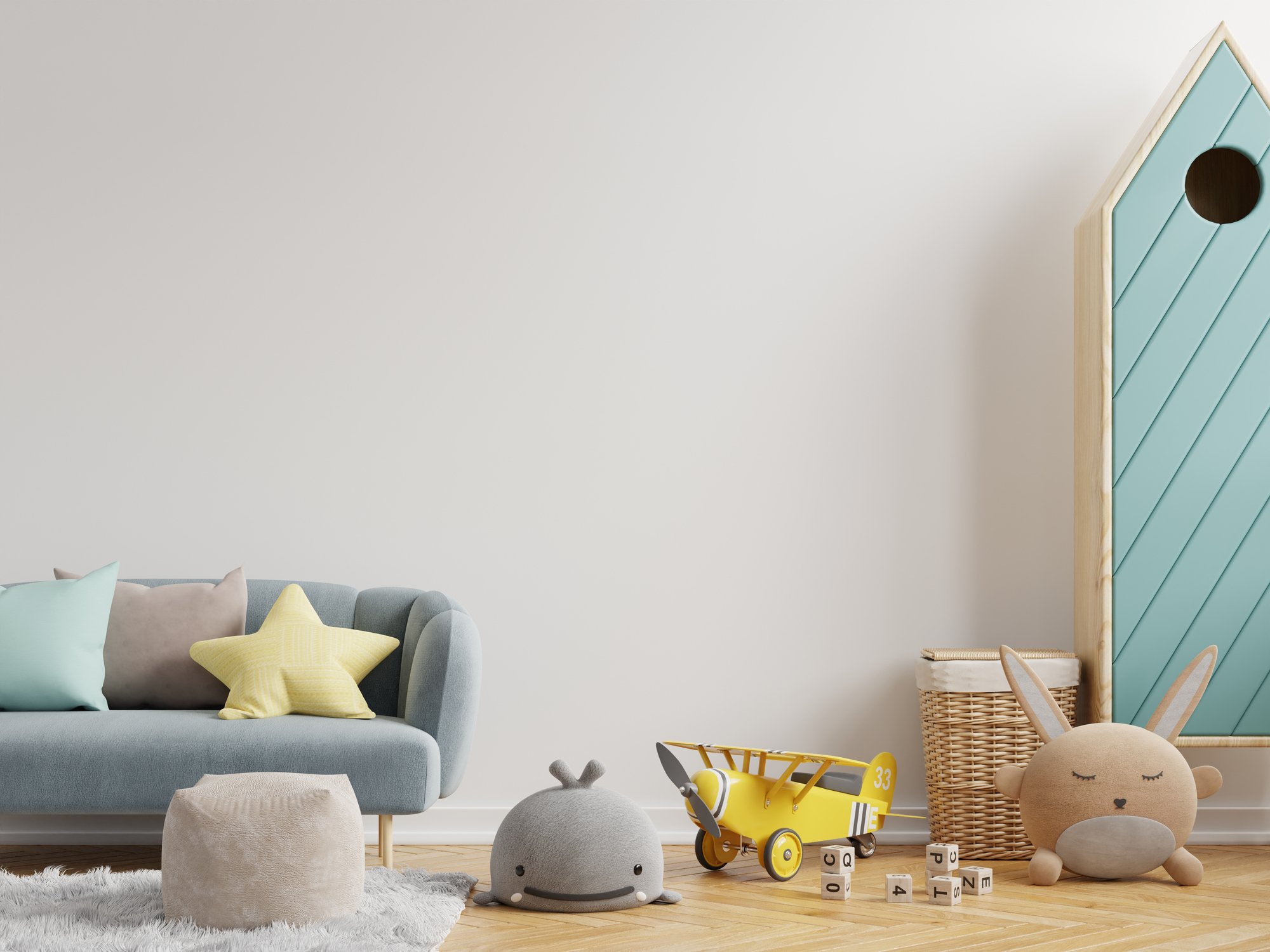 A children's playroom with a grey sofa and pastel-colored pillows, a white furry rug, plush and toy animals, a yellow toy airplane, a basket, and wooden blocks on a wooden floor. There's a pastel blue wooden playhouse wall panel on the right side.