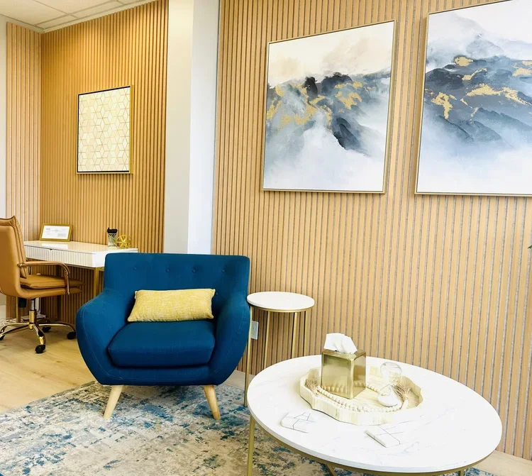 A cozy corner of a room with a blue armchair, a yellow pillow, a round white coffee table with tissue box and glass, a small side table, a desk with a beige chair, and two large abstract paintings on a wood-paneled wall.
