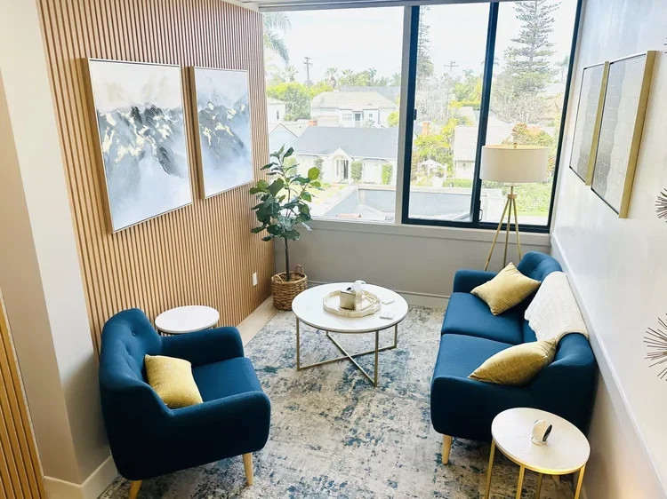 Cozy image of the Coronado Therapy Group office with two blue sofas, yellow pillows, a round coffee table, a floor lamp, a potted plant, and large windows showing outside.