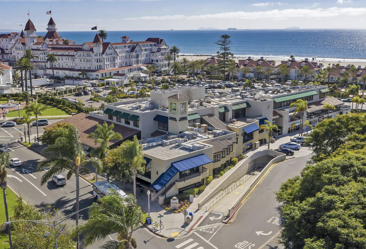 A coastal scene with a sandy beach and blue ocean in the background, and a beachfront hotel or resort with white architecture and red rooftops. There are palm trees, a parking lot, and streets with cars and bicycles in the foreground.
