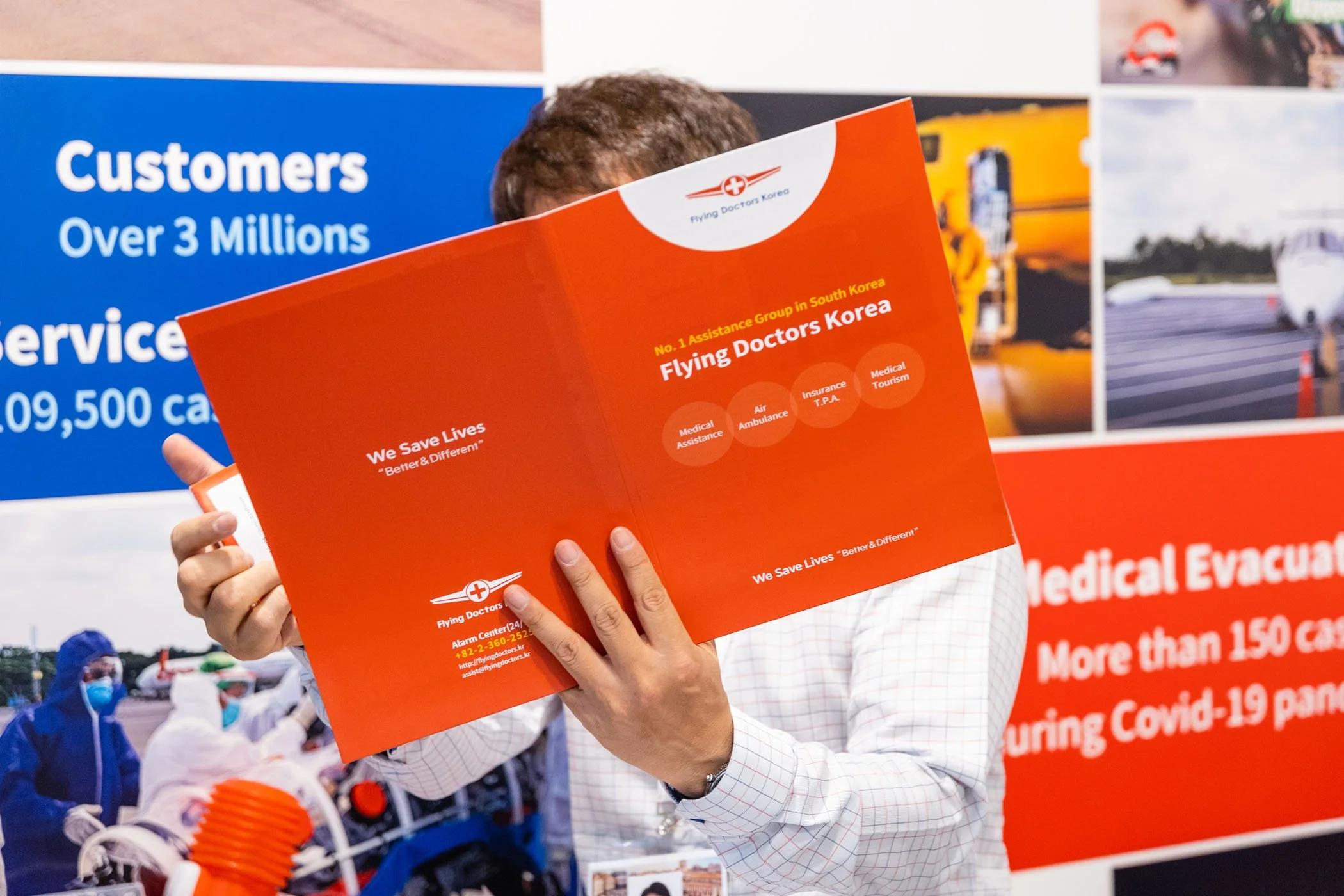 Persona leyendo folleto naranja que promociona los servicios de Flying Doctors Korea, incluyendo asistencia médica, ambulancia, seguro T.P.A. y turismo médico, en una exhibición con carteles y personal con mascarillas detrás.