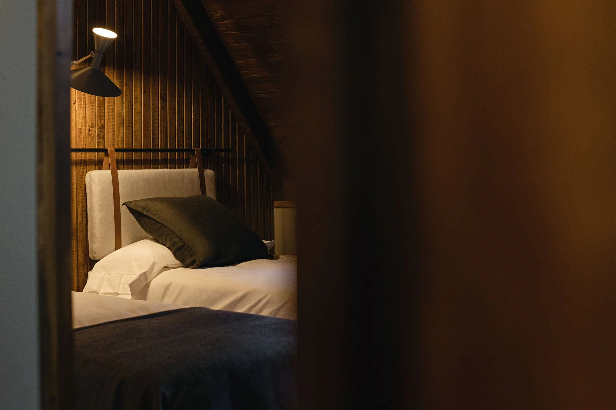 Cama en una habitación con paredes de madera, iluminada por una lámpara de lectura, con almohadas y ropa de cama en tonos neutros.