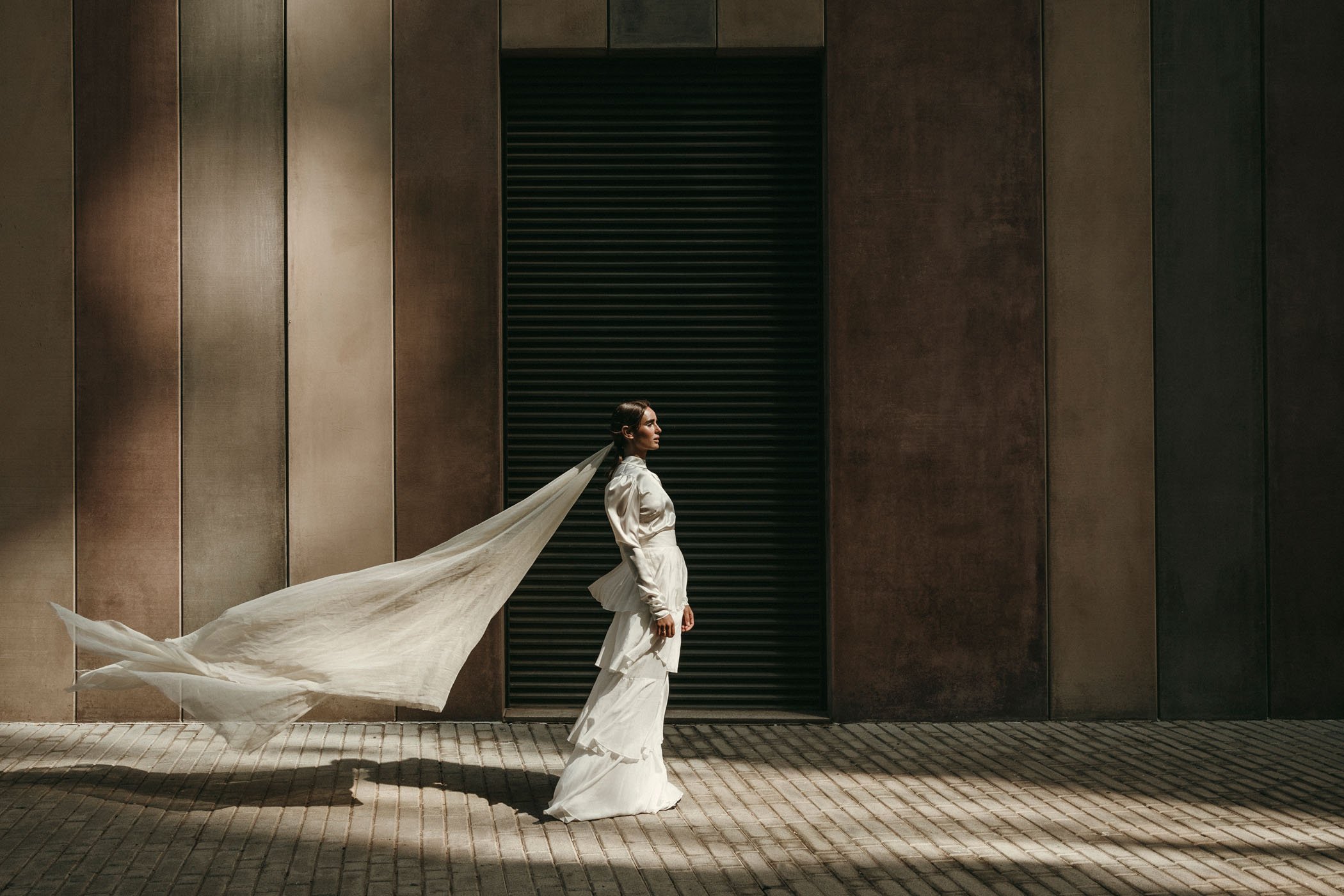 Mujer con vestido blanco y velo caminando en la calle con pared de fondo y sombra en el suelo
