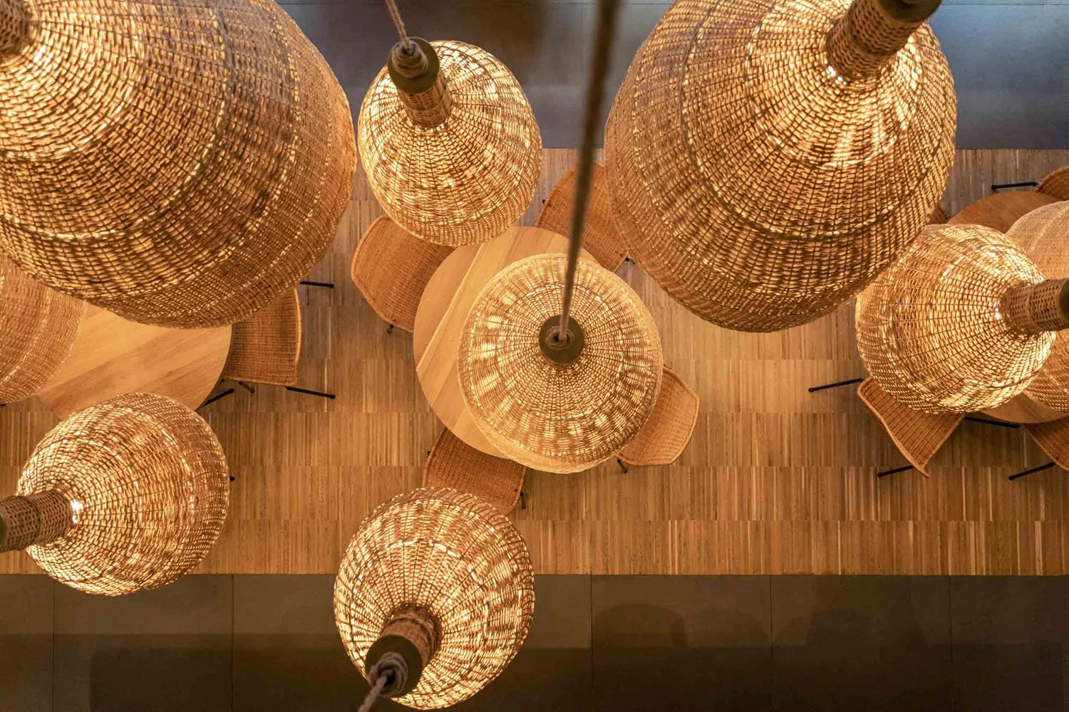 Lámparas de mimbre colgadas del techo, vista desde abajo, sobre mesas de madera y sillas con asiento de madera.