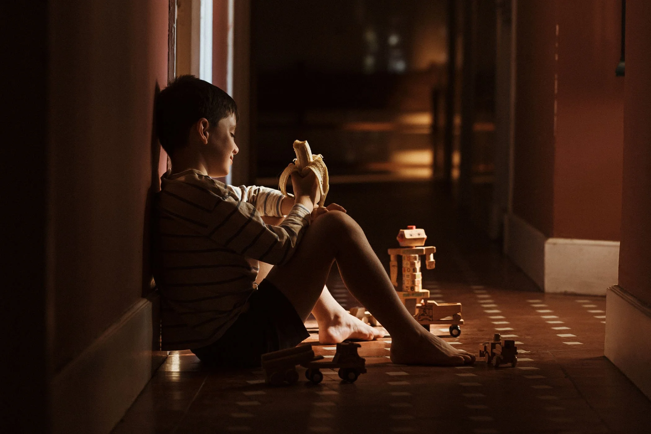 Niño sentado en el suelo de un pasillo jugando con juguetes de madera y comiendo un plátano, iluminación cálida y ambiente tranquilo.