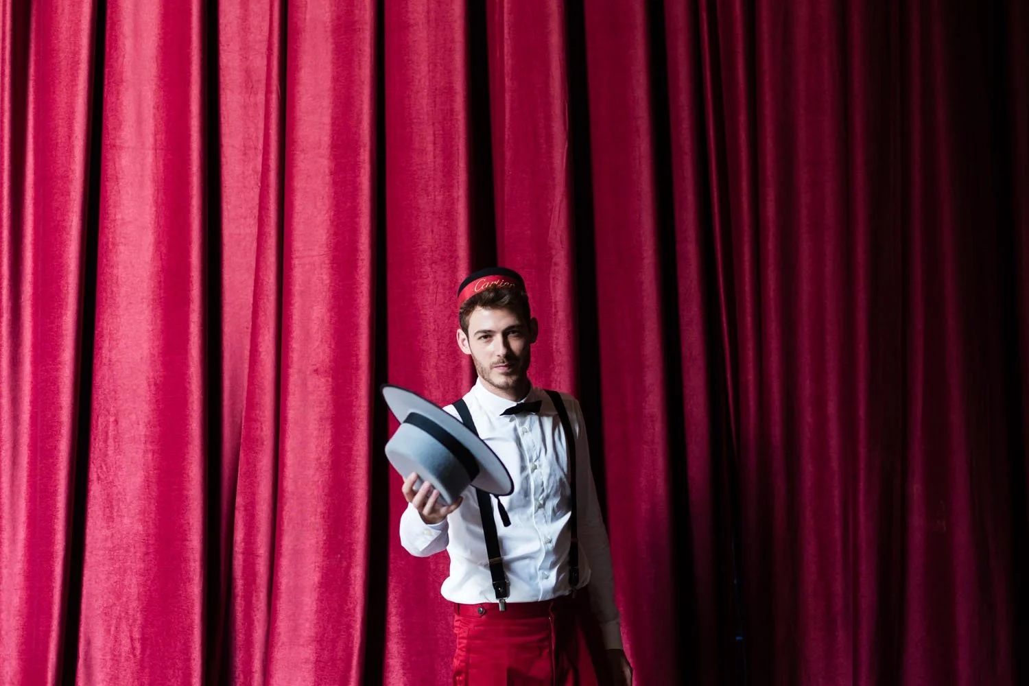 Hombre vestido de showman con sombrero y ropa formal, sosteniendo un sombrero, frente a cortinas rojas del teatro.