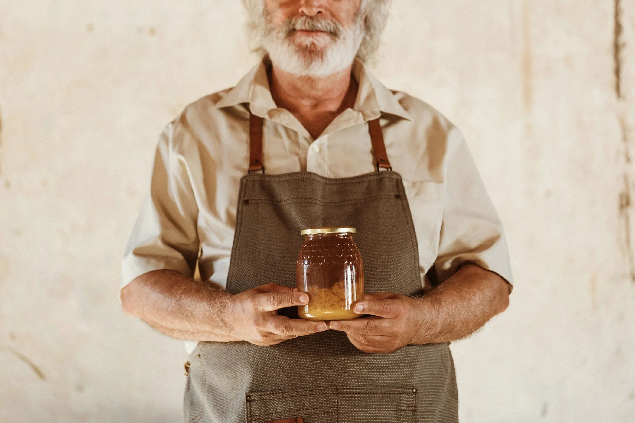 Hombre de cabello canoso y barba, sosteniendo un frasco de miel con ambas manos, lleva una camiseta beige y un delantal marrón en un fondo de pared clara y texturizada.