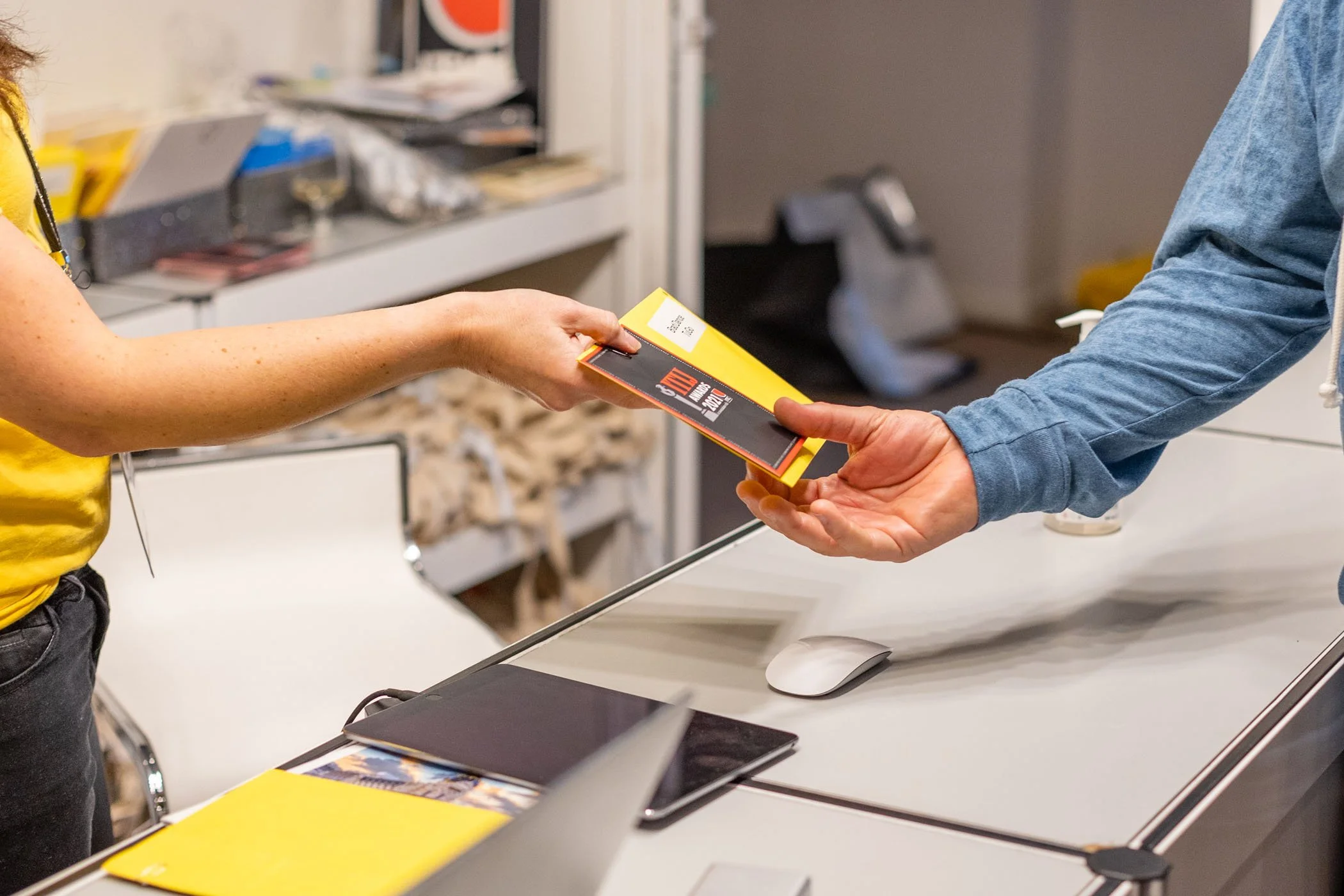 Una persona entregando un paquete a otra en una oficina, la escena muestra una mesa con computadora y varios objetos en el fondo.