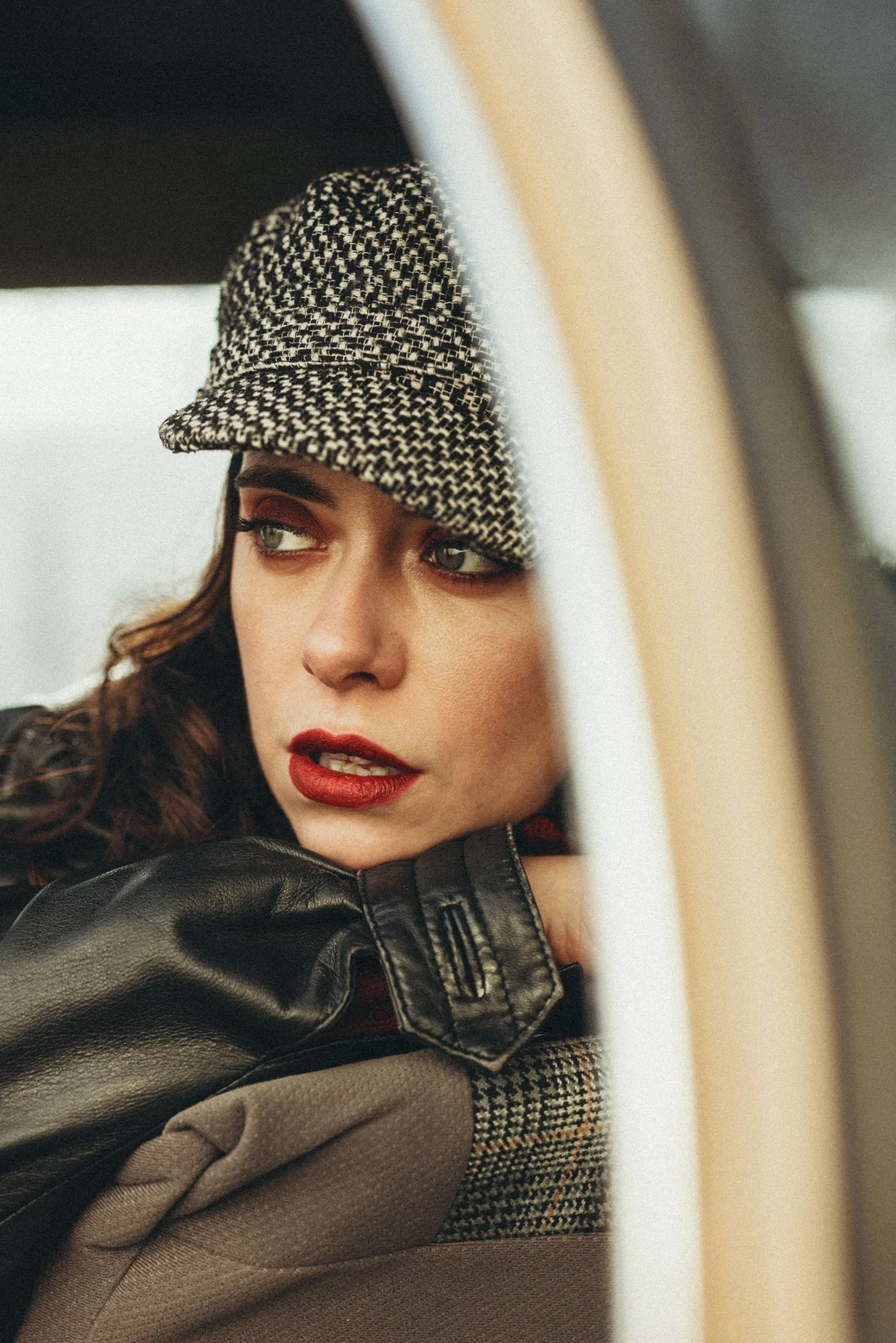 Retrato de una mujer con gorra de mezclilla, guantes de cuero y una chaqueta con detalles de tela a cuadros, mirando pensativa a través de la ventana.