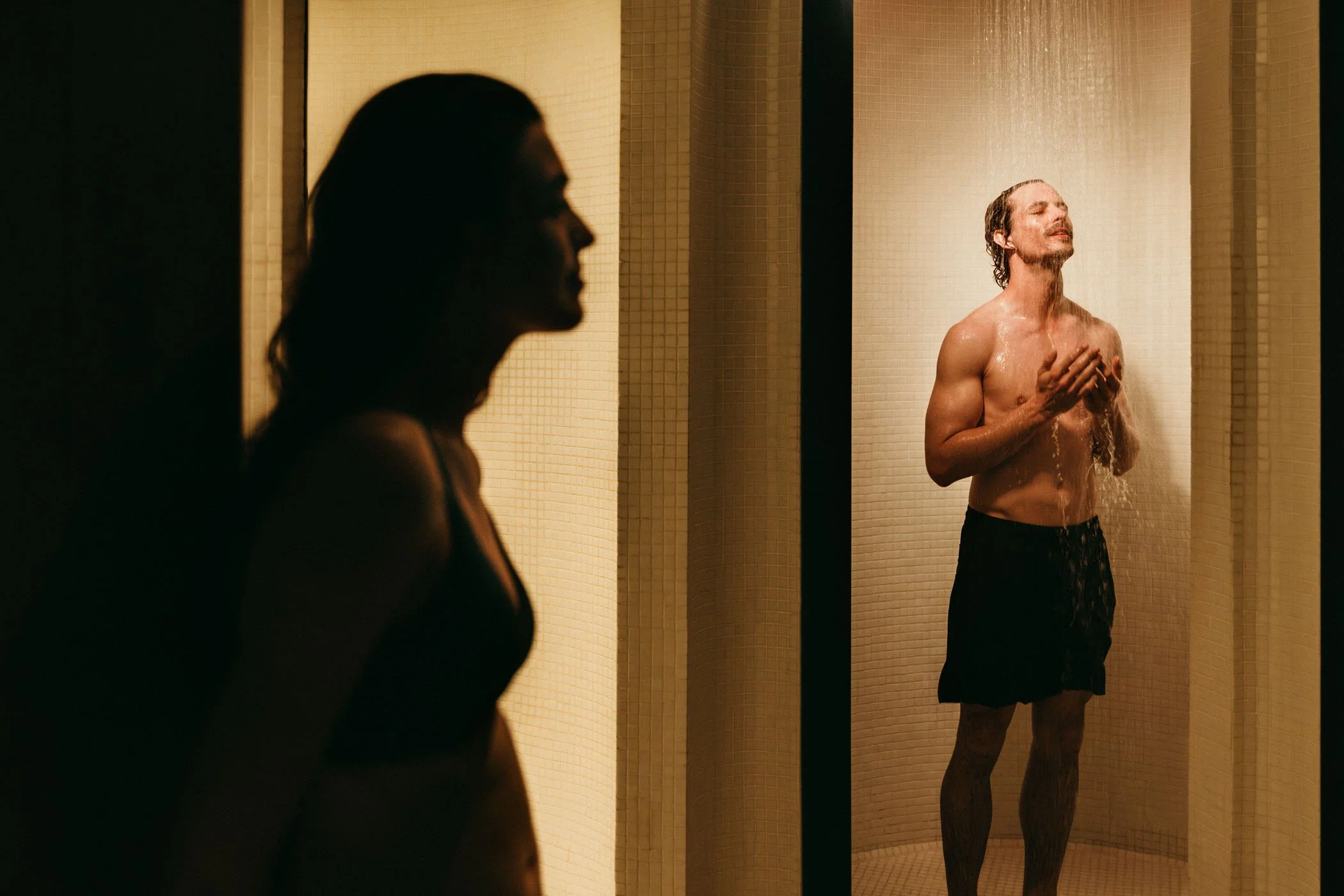 Dos personas en una ducha, una mujer en la esquina izquierda en sombra y un hombre en la esquina derecha, recibiendo una ducha de agua caliente.