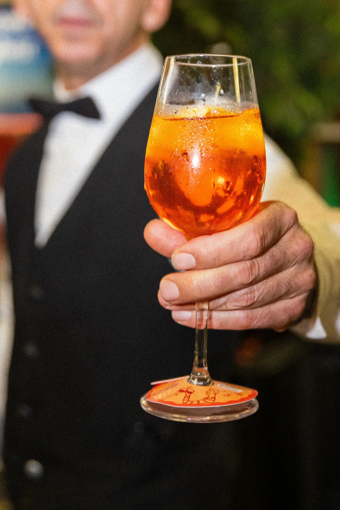 Una persona sosteniendo una copa de cóctel color naranja con hielo, con un fondo desenfocado y una iluminación cálida.