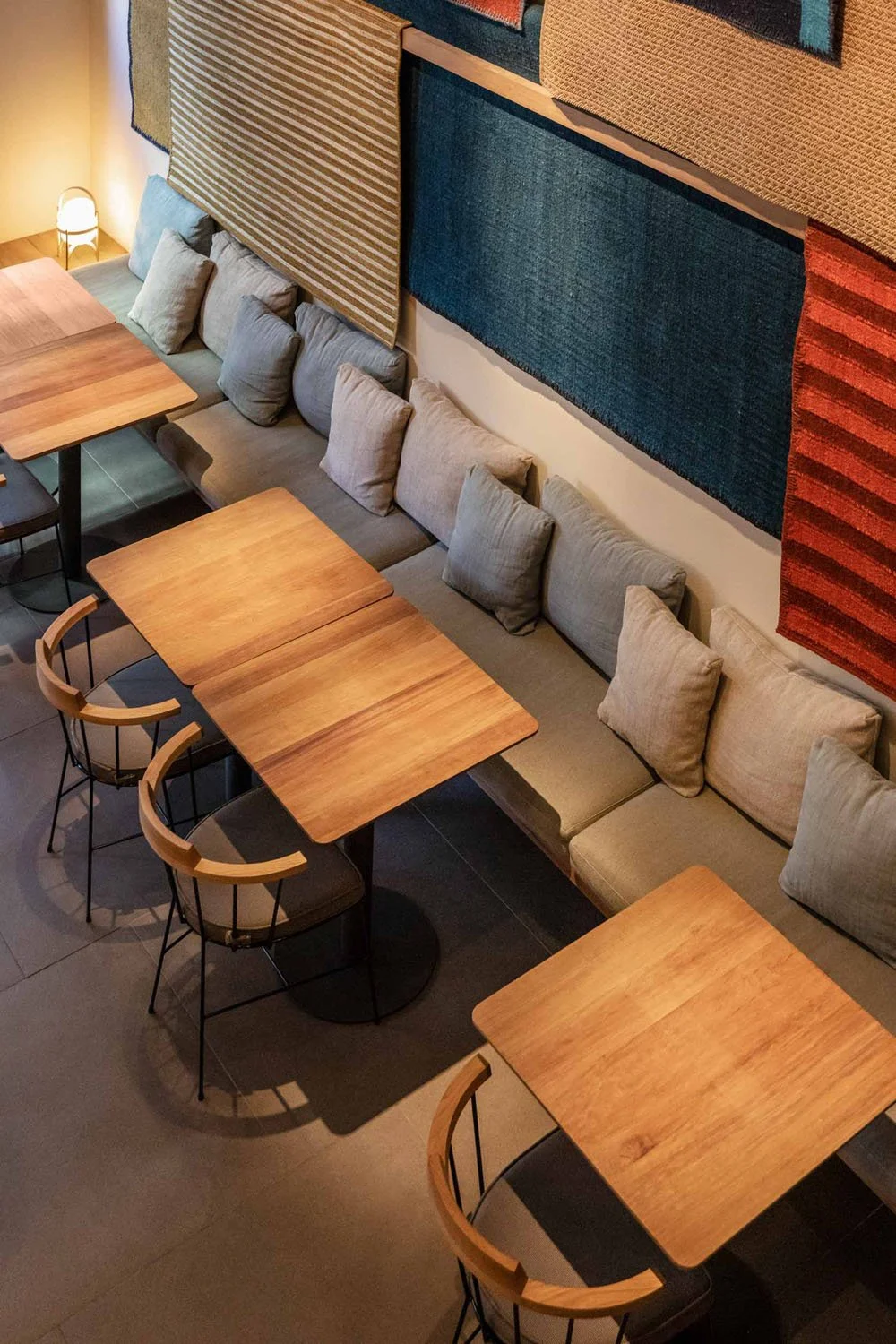 Vista aérea de una cafetería con mesas de madera y sillas, paredes decoradas con tapices o alfombras en diferentes colores y texturas.