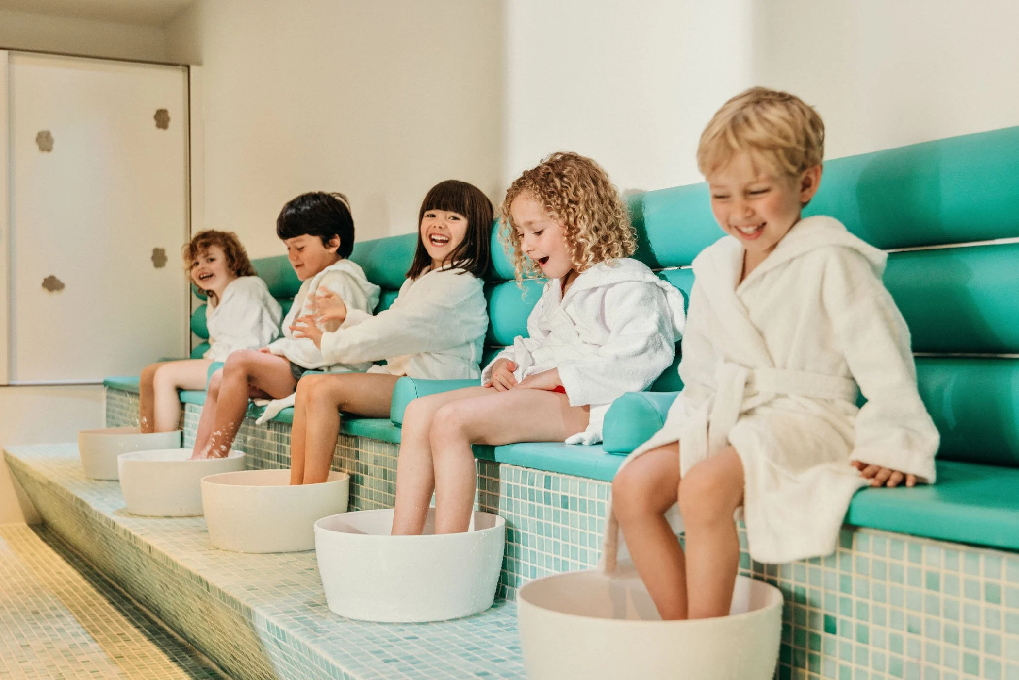 Niños con toallas blancas disfrutando de un baño en un spa o piscina, con los pies en cubetas y tarros de lavado, riendo y hablando mientras se secan.