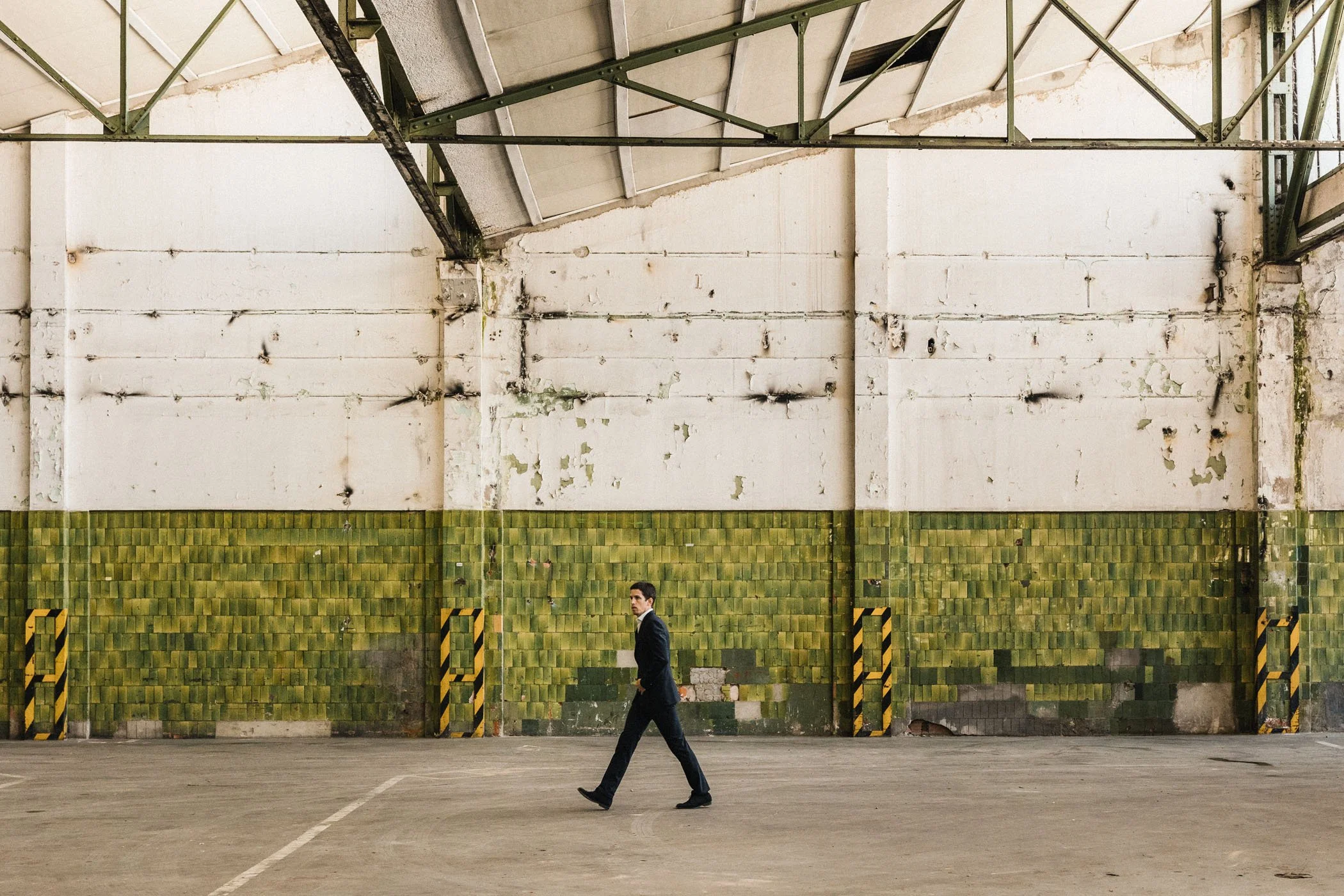 Hombre de traje negro caminando en un espacio industrial vacío con paredes de bloques verdes y techo de metal