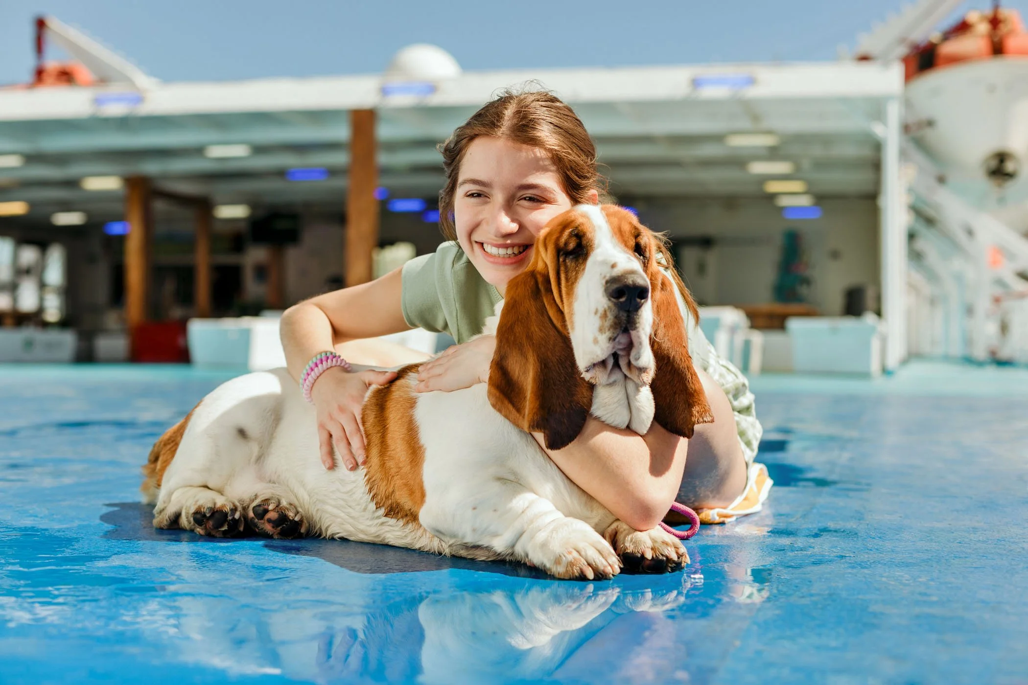 Una niña feliz abrazando a un perro beagle en una piscina cubierta, con fondo de estructura de madera y agua azul.