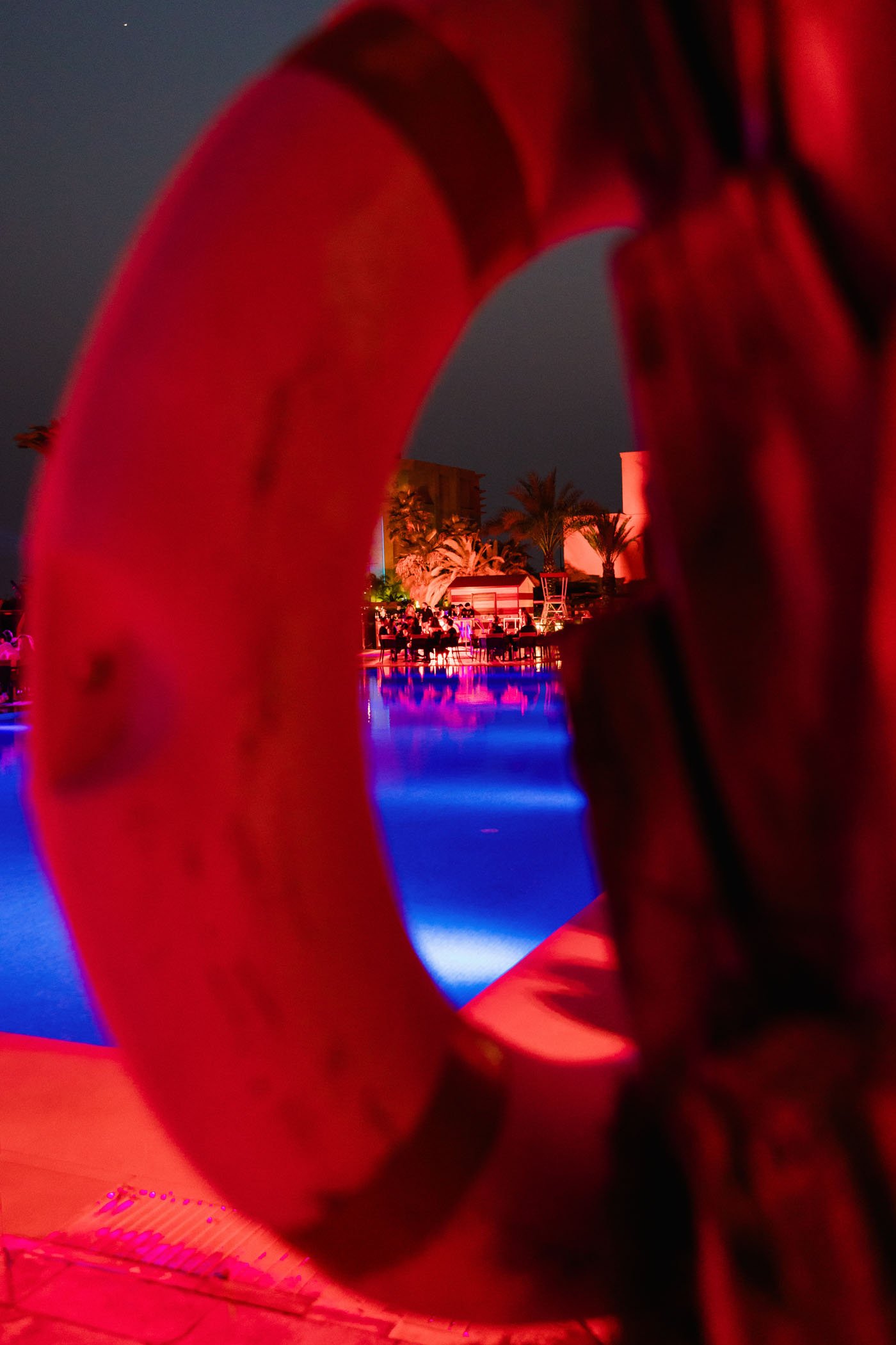 Junto a una piscina iluminada por luces de colores, se observa una figura de un cinturón salvavidas en primer plano, con una escena nocturna de una fiesta o reunión social con palmeras y personas en el fondo.