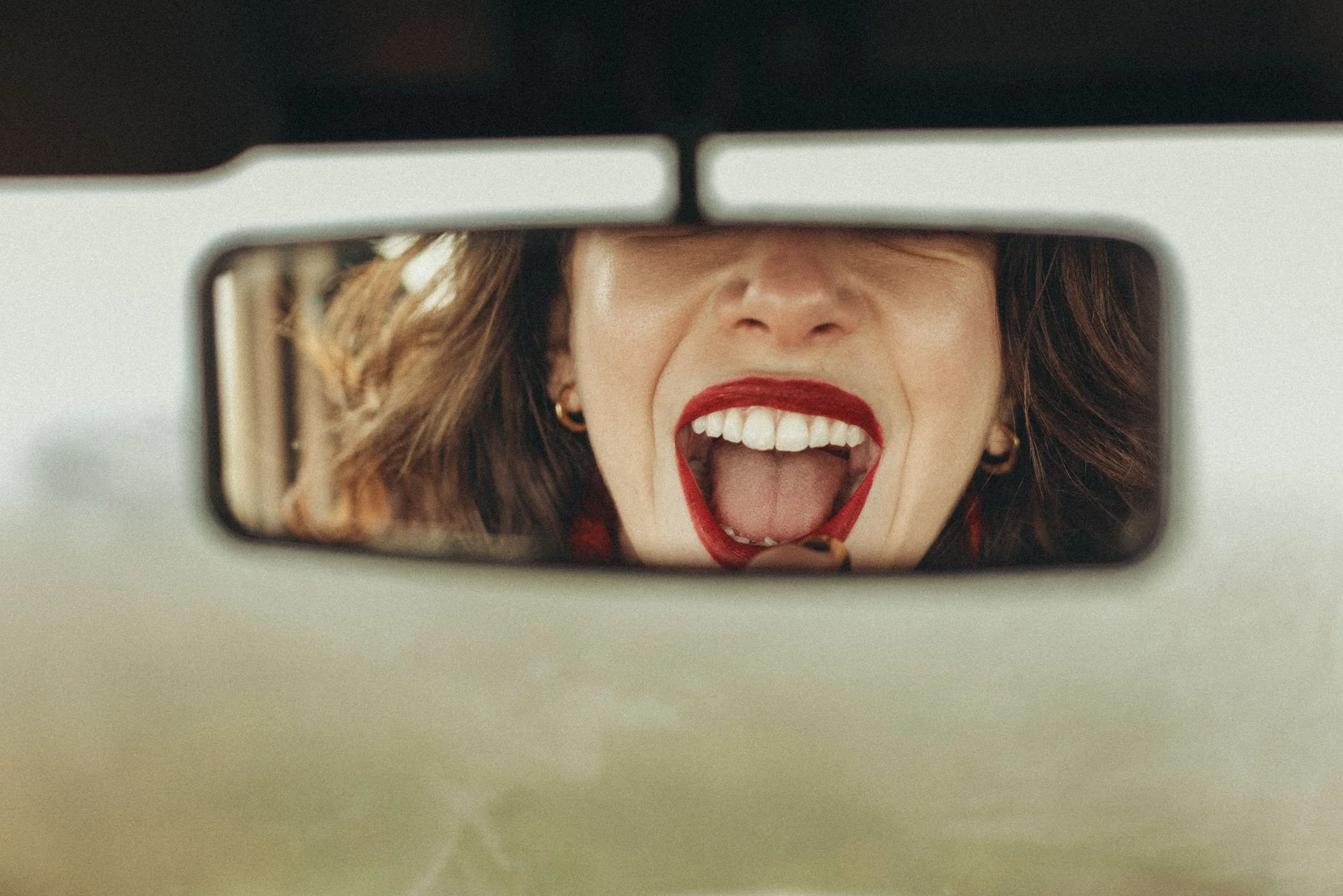 Mujer con labios rojos y lentes de contacto, abriendo la boca y haciendo una expresión de sorpresa o diversión, vista en el espejo del espejo retrovisor del automóvil.