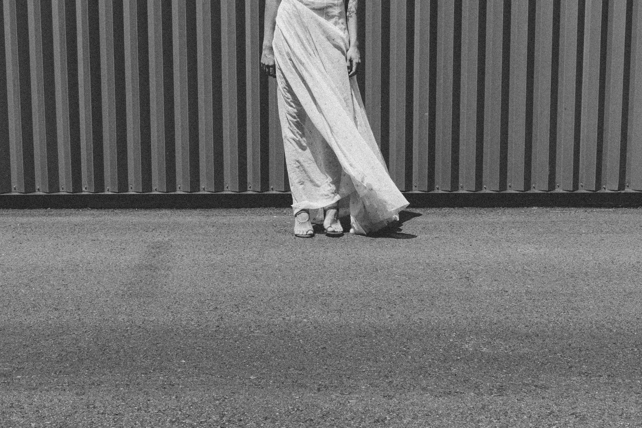 Persona de espaldas con pantalones anchos y tacones altos, de pie frente a una pared de metal.