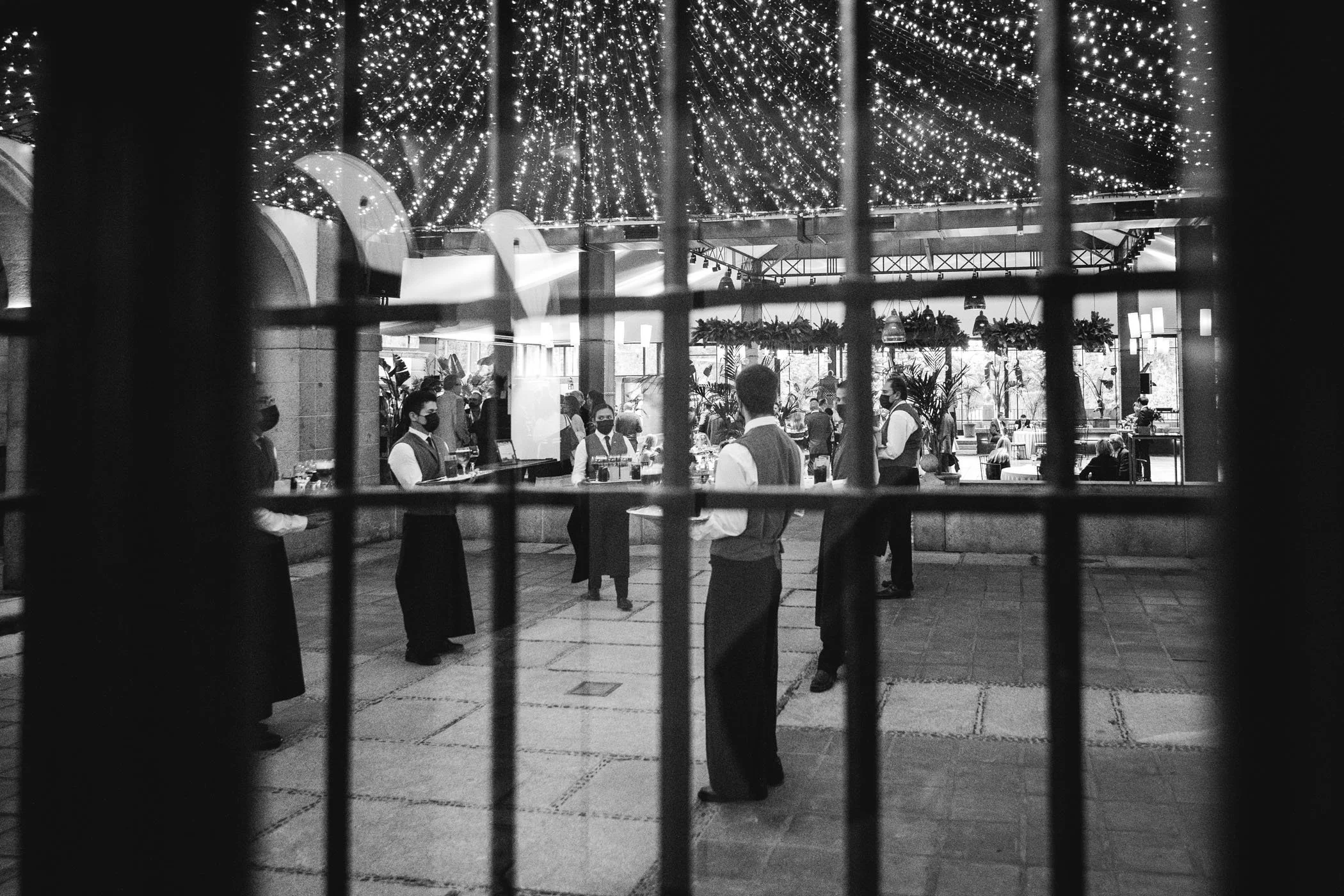 Vista de un restaurante elegante con músicos tocando en un ambiente decorado con luces navideñas, vista a través de una reja negra.