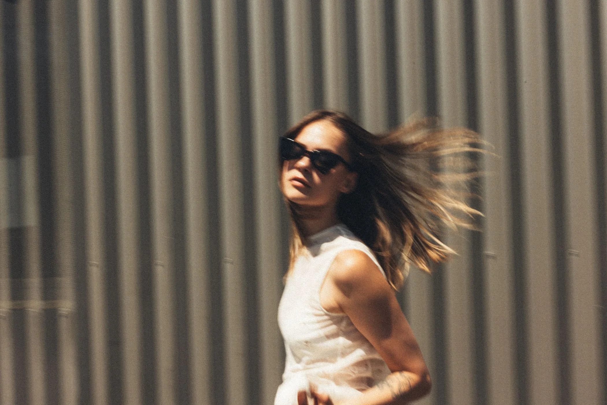 Mujer con gafas de sol, vestido blanco, caminando frente a una pared de metal vertical, con el cabello al viento.