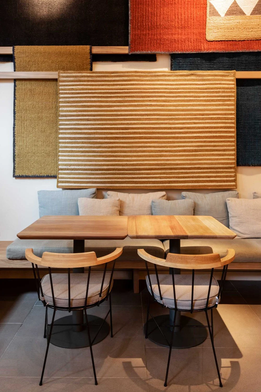Muebles de cafetería con mesa de madera, sillas de metal con cojines, sillones con cojines y tapices decorativos en la pared con diferentes texturas y colores.