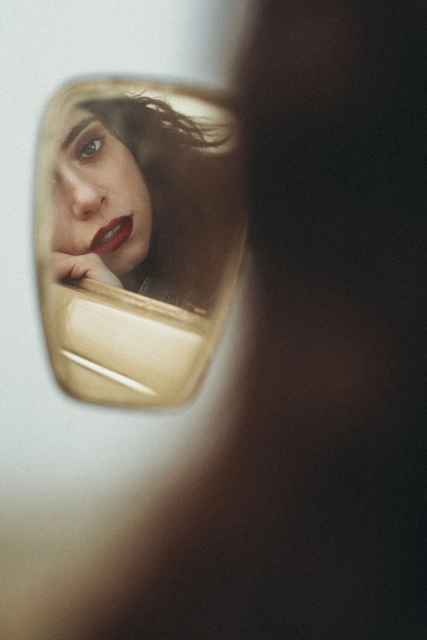 Joven mujer con maquillaje oscuro, rojo de labios, y cabello rizado se mira en un espejo, reflejando su rostro y expresión pensativa.