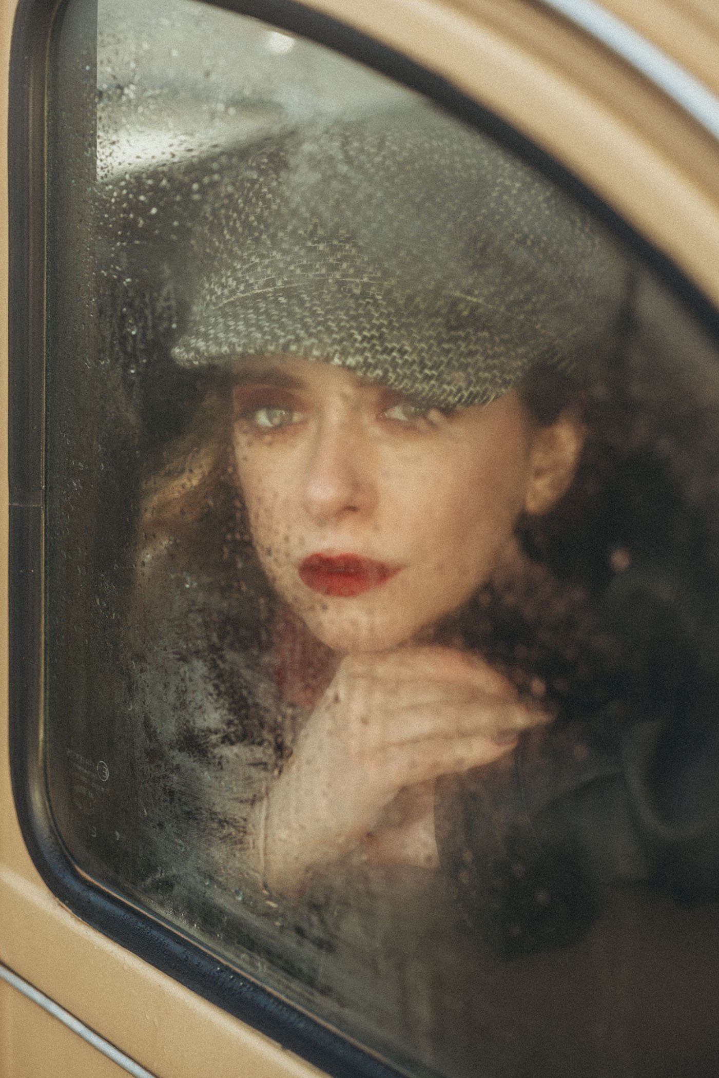 Mujer mirando por la ventana empañada con expresión pensativa, lleva un sombrero de cuadros y tiene el cabello rizado.