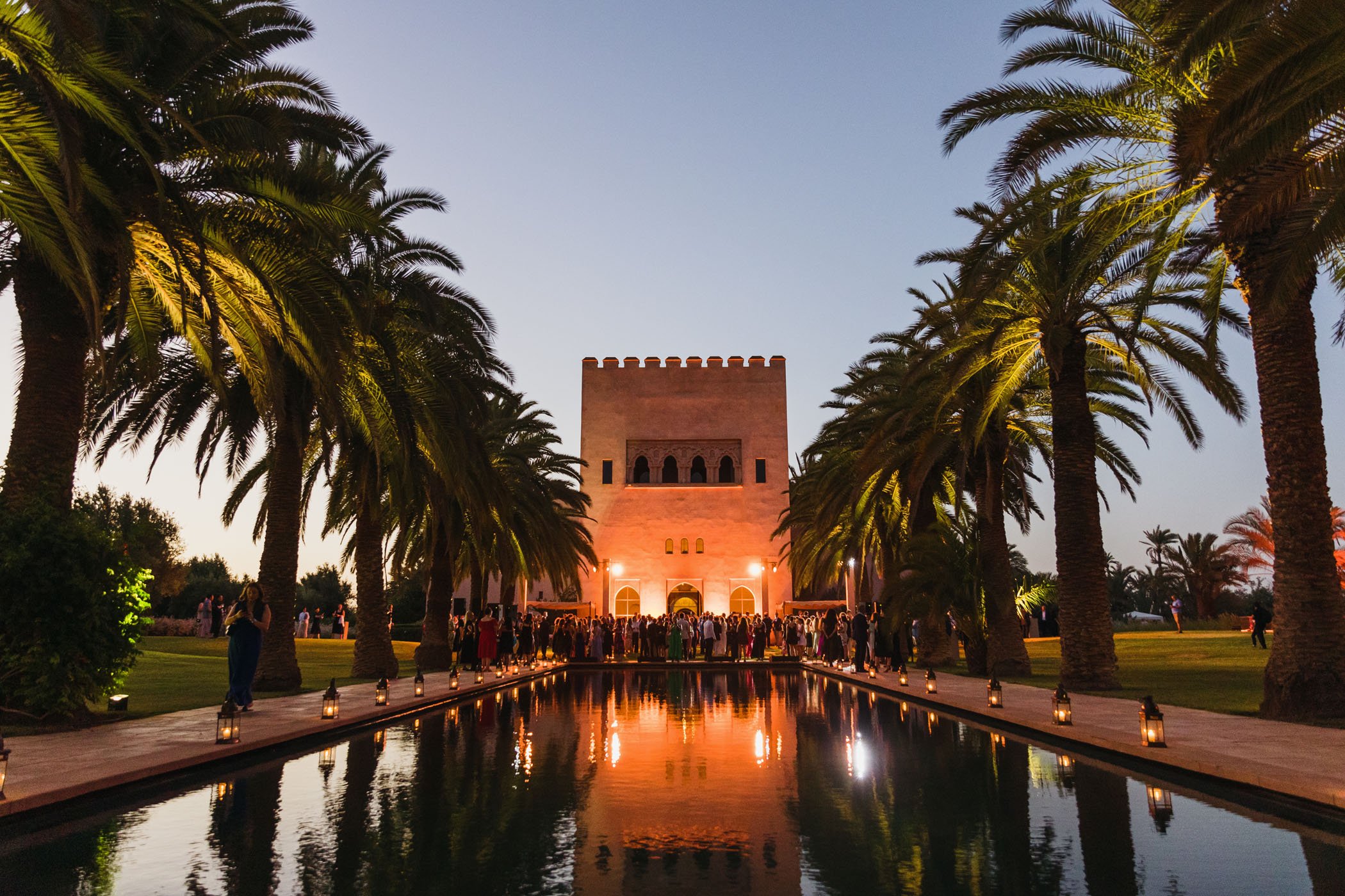 Vista de un paseo con un estanque reflejando un edificio estilo fortaleza y palmeras a los lados, con personas en una evento al atardecer.