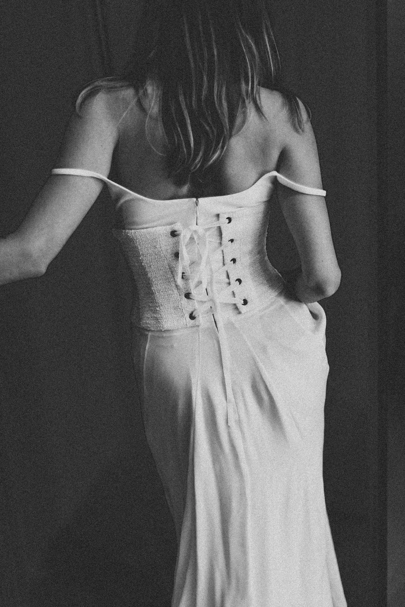 Mujer con vestido de tirantes y corset ajustado, de espaldas, en un entorno oscuro, en una foto en blanco y negro.