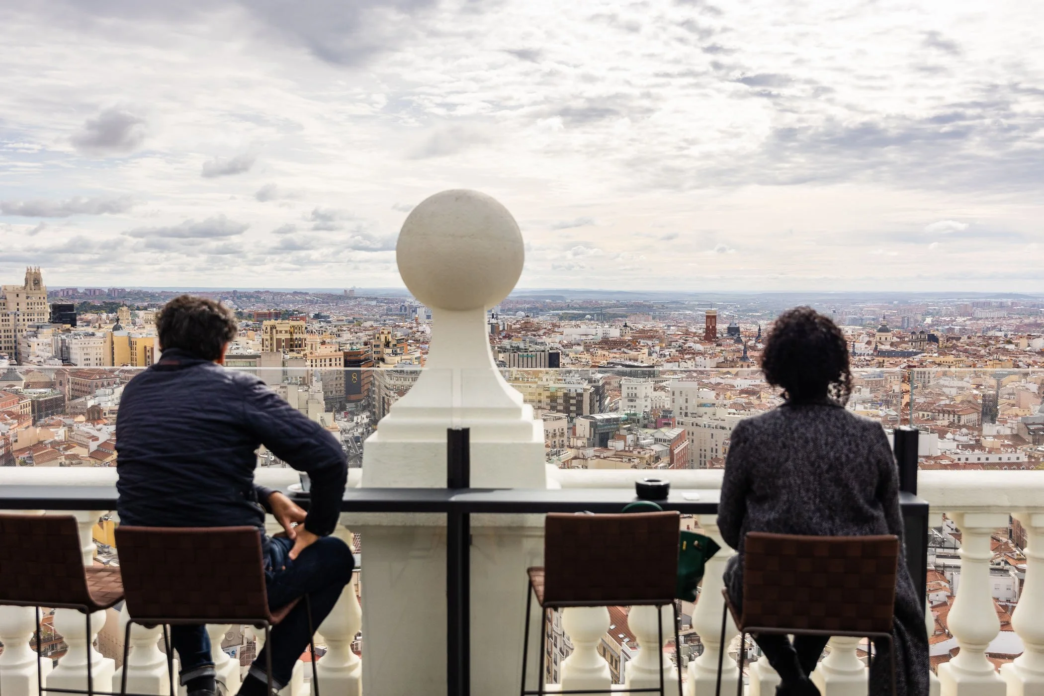 Dos personas sentadas en un mirador con vista panorámica de una ciudad, en un día nublado.