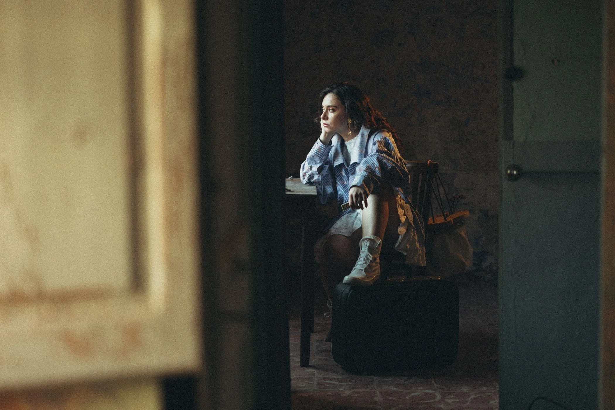 Una mujer joven con cabello oscuro y rizado, sentada en una silla, pensativa, descansando su rostro en su mano, en un ambiente oscuro y con luz suave, dentro de una habitación con paredes de piedra y una mesa al fondo con algunos objetos.
