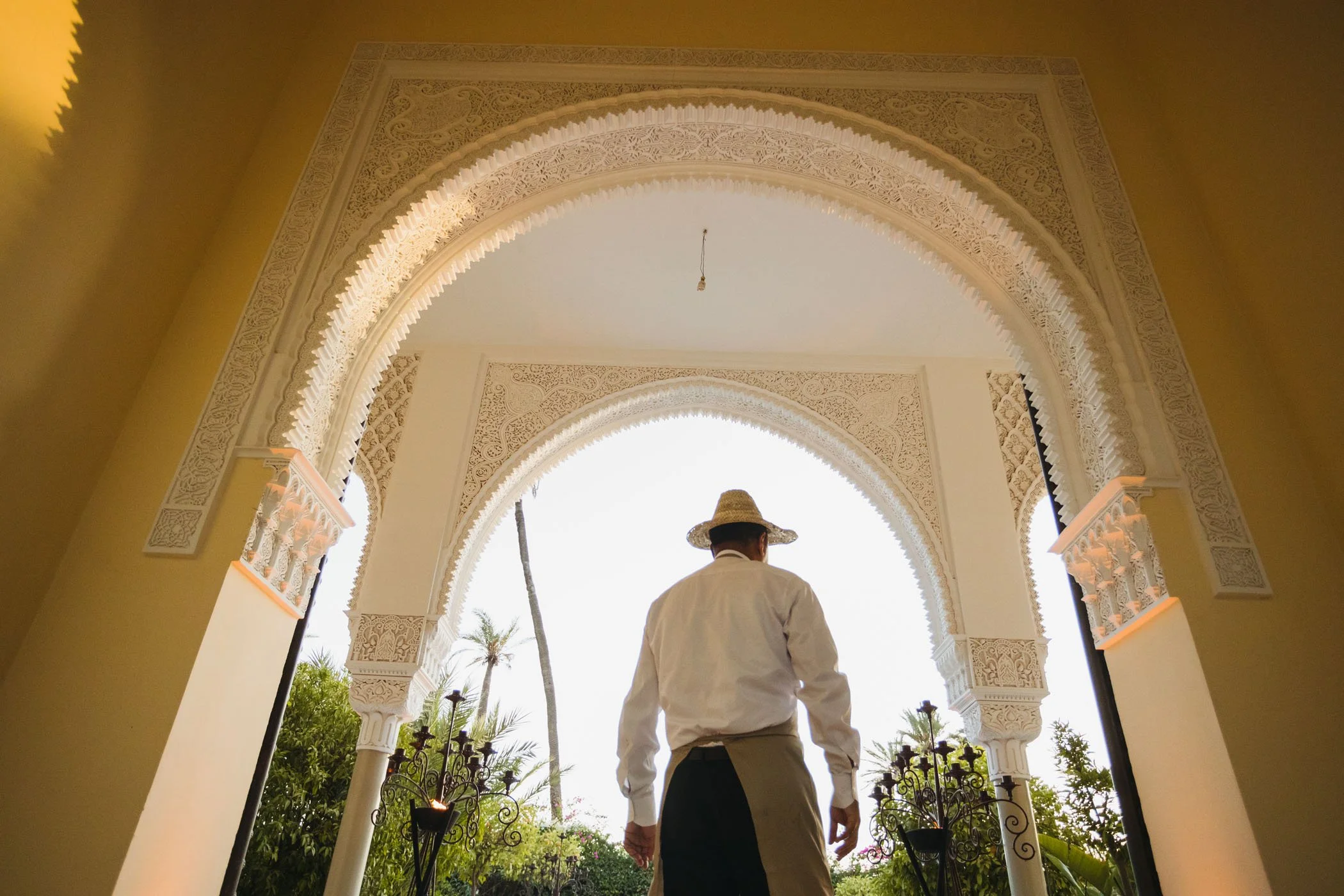 Hombre con sombrero tradicional mexicano de espiga, vestido con camisa blanca y delantal oscuro en entrada de arcos decorativos, con plantas y lantinas en el fondo.