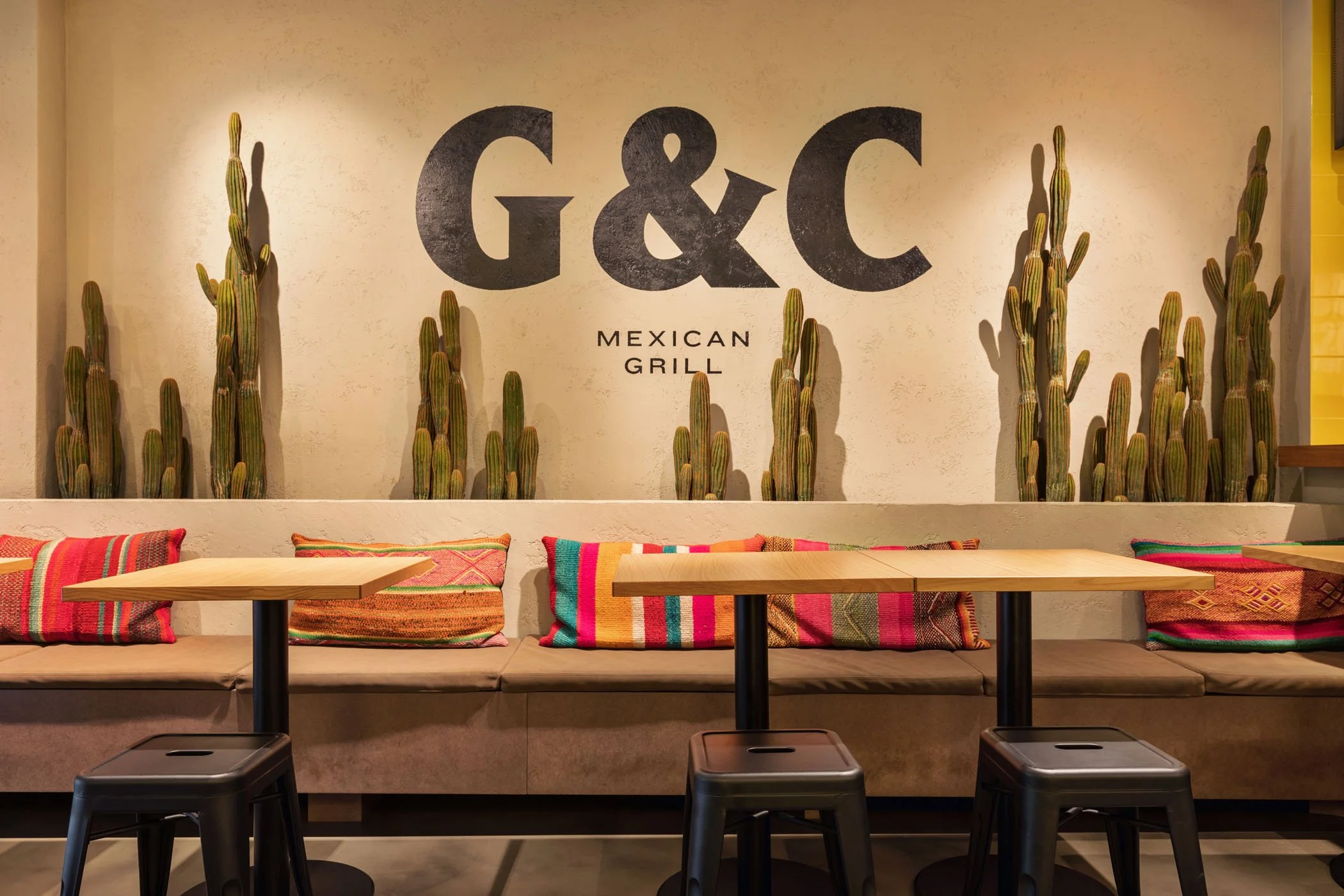 Interior de restaurante G&C Mexican Grill con sillas, mesas de madera y cojines coloridos, plantes de cactus decorativos en la pared.