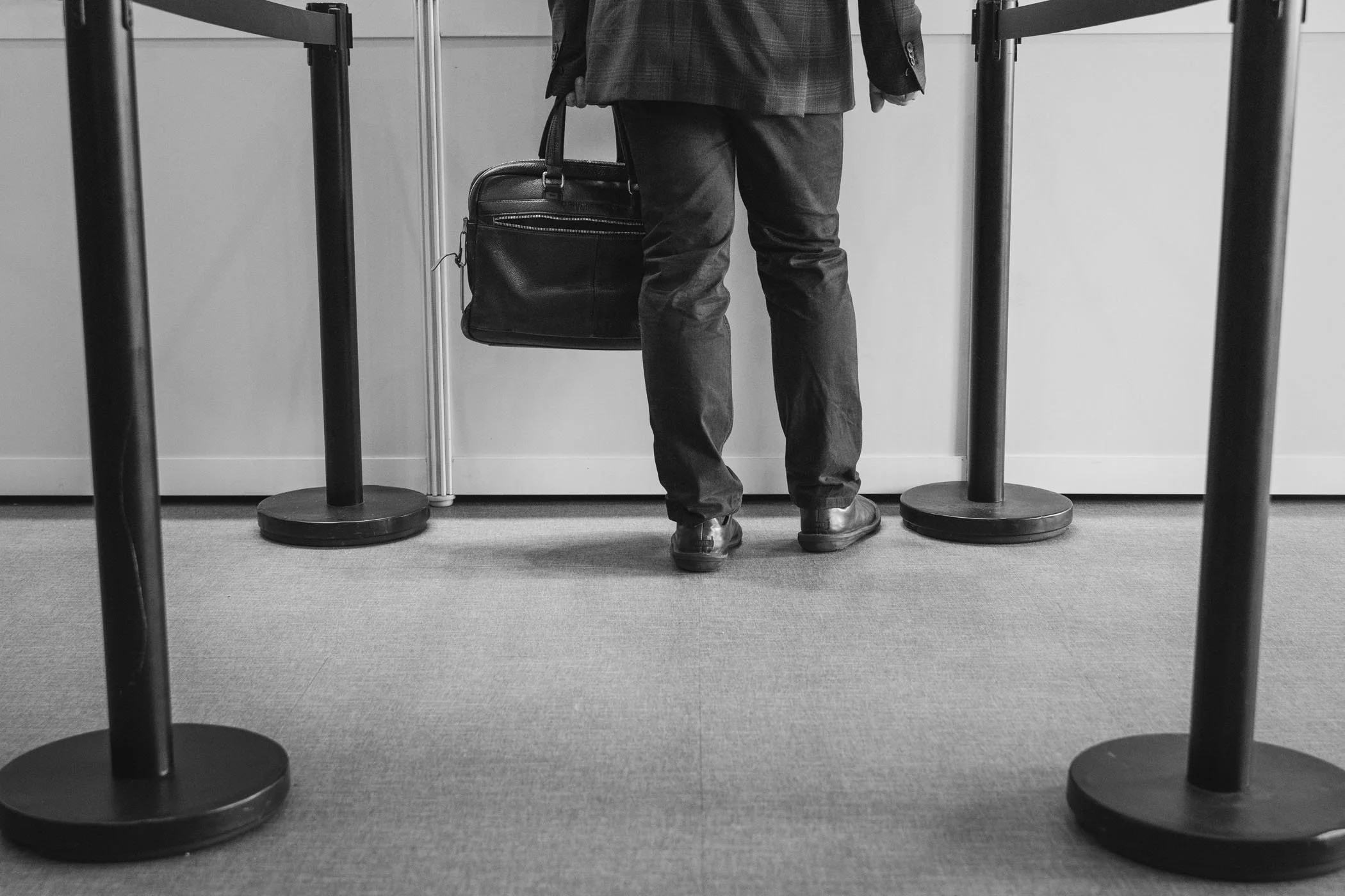 Persona de espaldas en fila con maletín, esperando en una fila con barreras en un espacio interior.