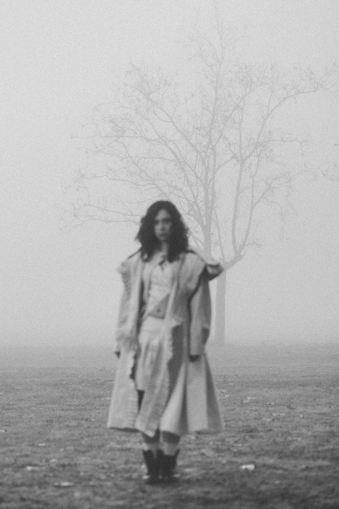Mujer de cabello rizado y vestimenta larga caminando en un campo con la niebla y un árbol desnudo en el fondo en una foto en blanco y negro.