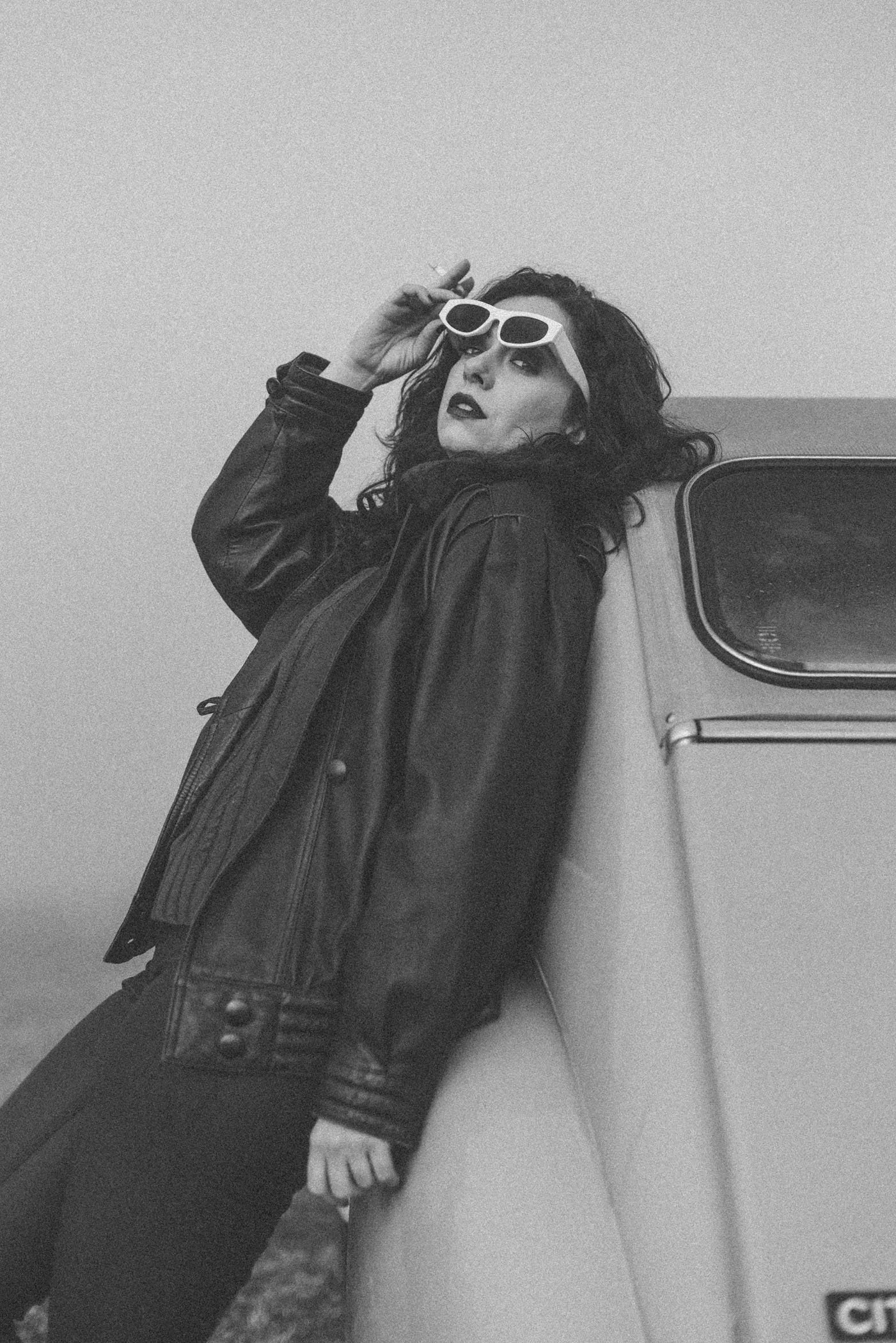 Mujer con gafas blancas, chaqueta de cuero, apoyada en un vehículo, en fotografía en blanco y negro.