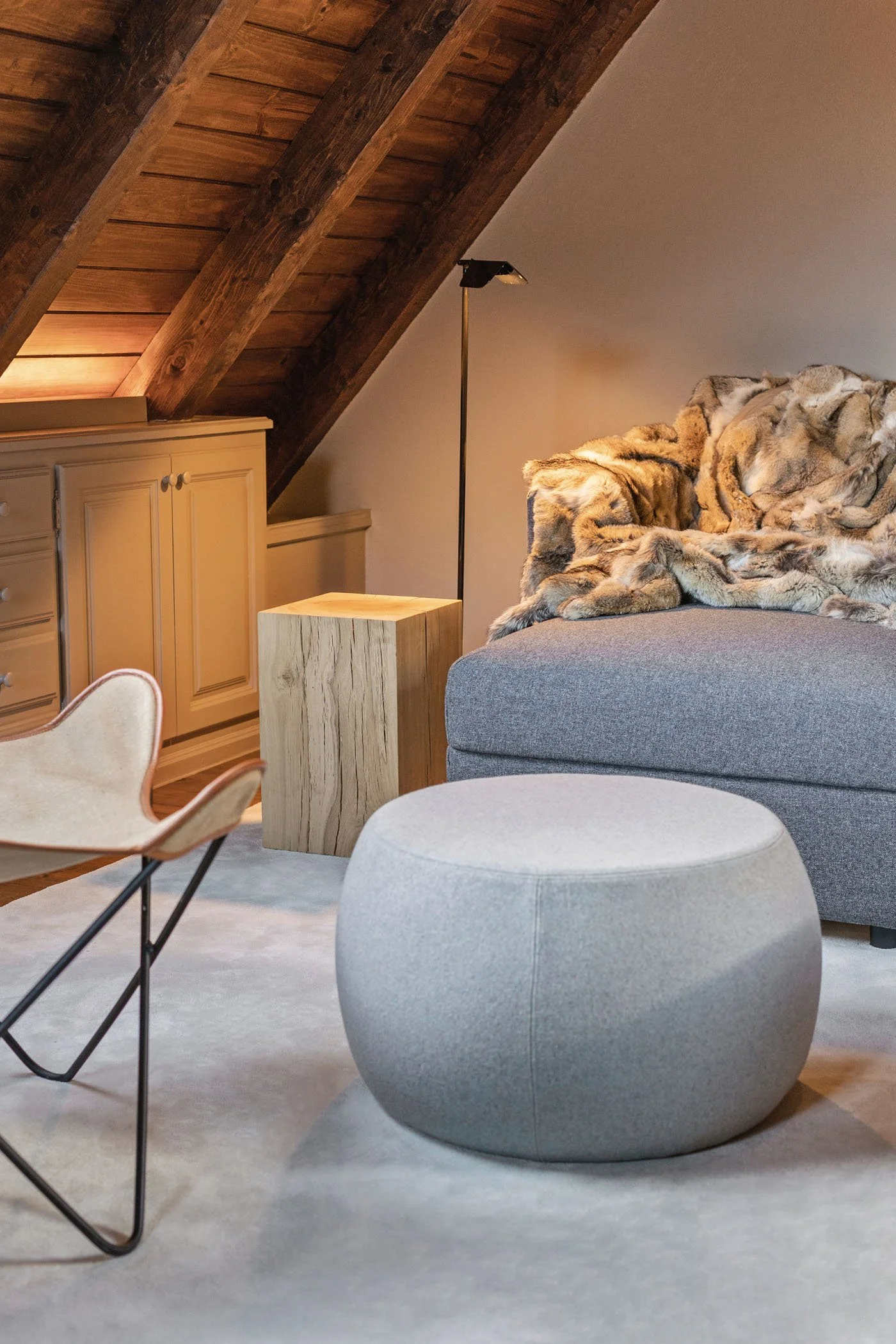 Sala de estar en un ático con muebles de tonos neutros, una lámpara delgada, un sofá con cubierta de piel, un puff y un armario de madera, con techos de madera en pendiente.