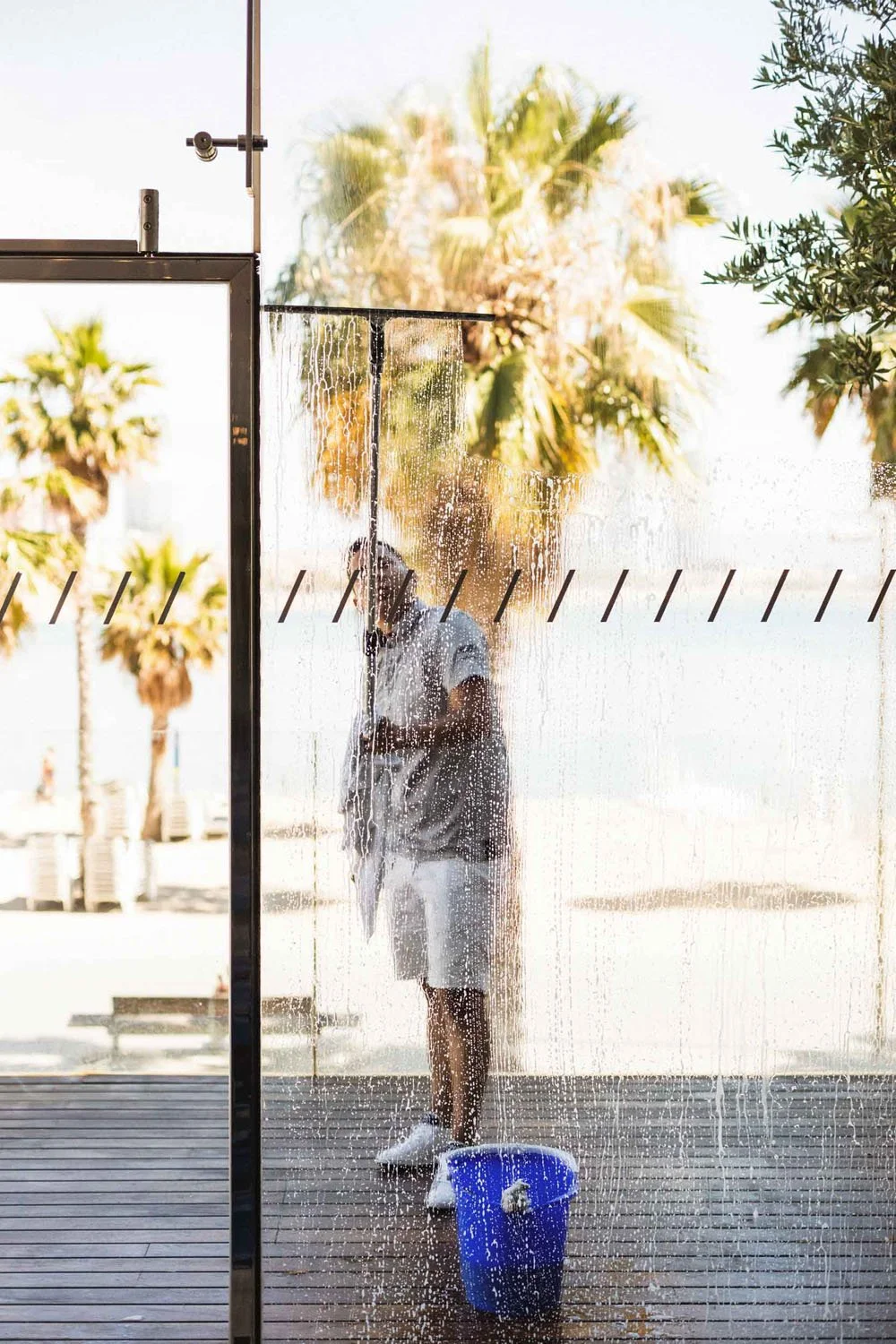 Persona limpiando una mampara de vidrio exterior, con árboles de fondo y un cubo azul en el suelo.