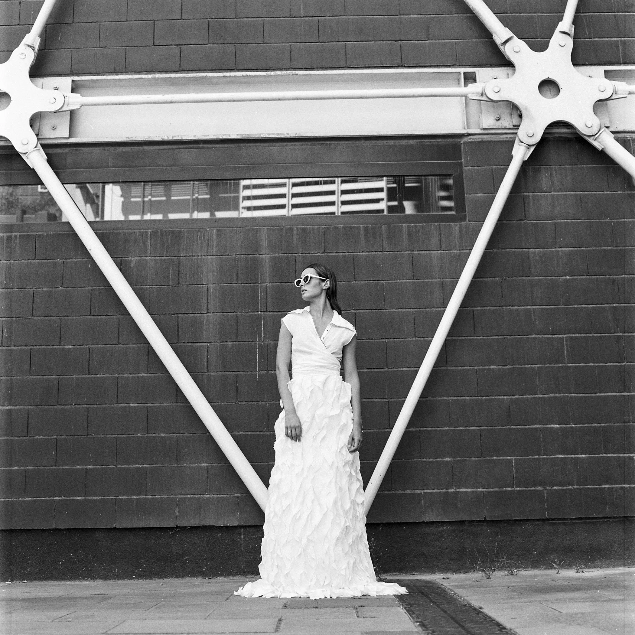 Mujer vestida con un vestido largo blanco y gafas de sol blancas, de pie frente a una pared de ladrillos en una ciudad, en una imagen en blanco y negro.