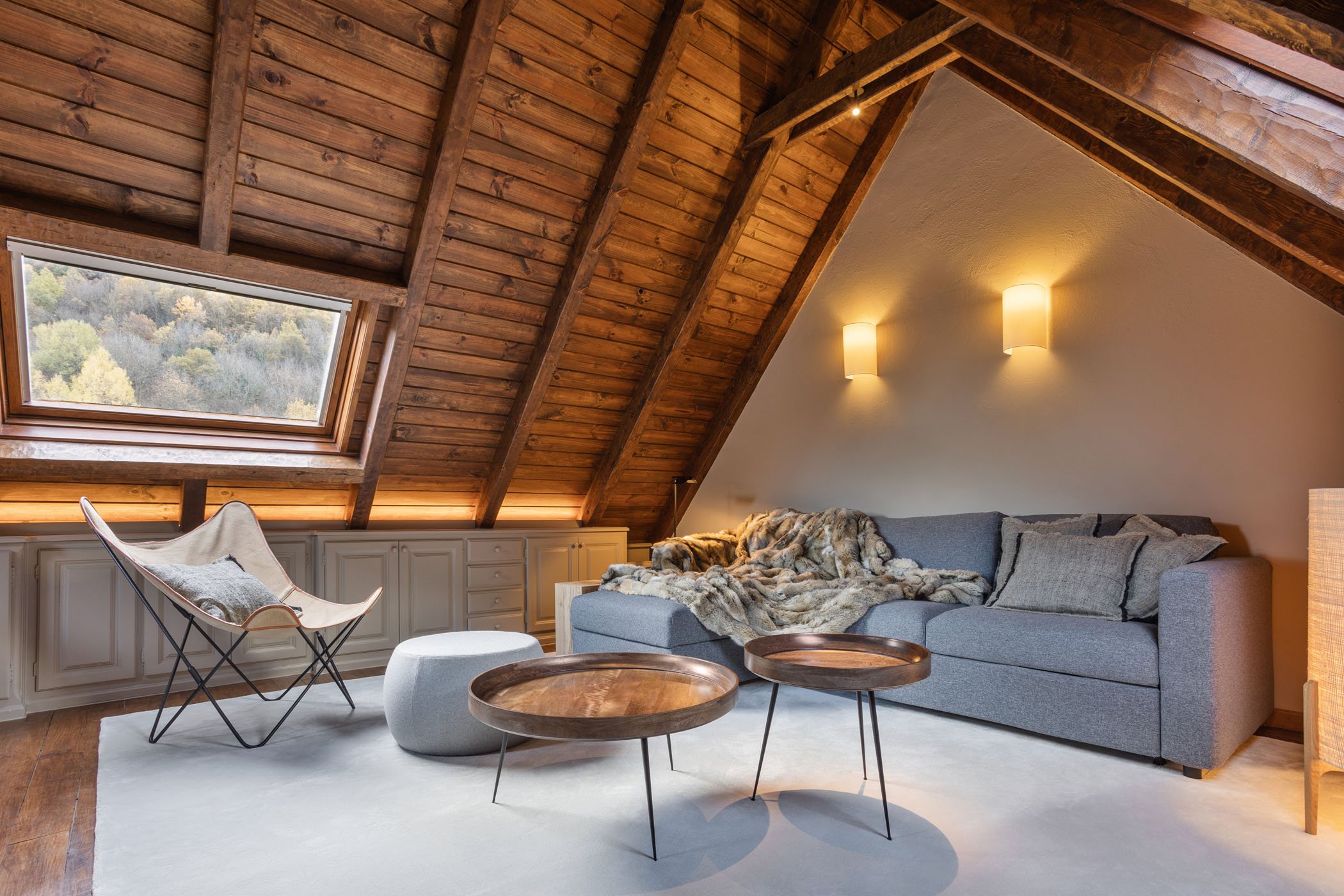 Sala de estar en un ático con paredes y techo de madera, una ventana panorámica que muestra árboles, un sofá gris con una manta de piel, mesas de centro de madera, una silla tipo cuna y un sillón gris, iluminación cálida en las paredes y una alfombra