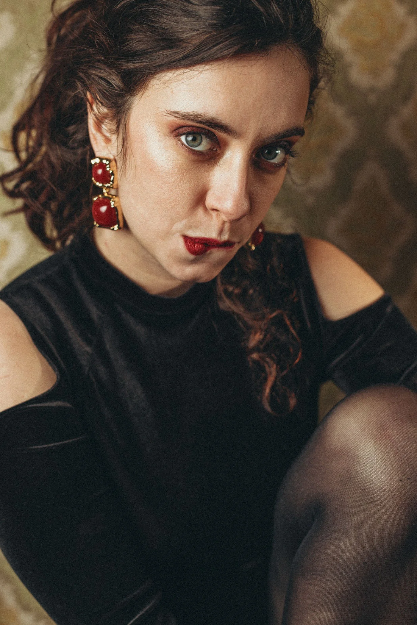 Retrato de una mujer con cabello rizado y ojos claros, usando un vestido negro con hombros descubiertos y pendientes grandes rojos, con maquillaje rojo en los labios, en un entorno de fondo oscuro y rugoso.