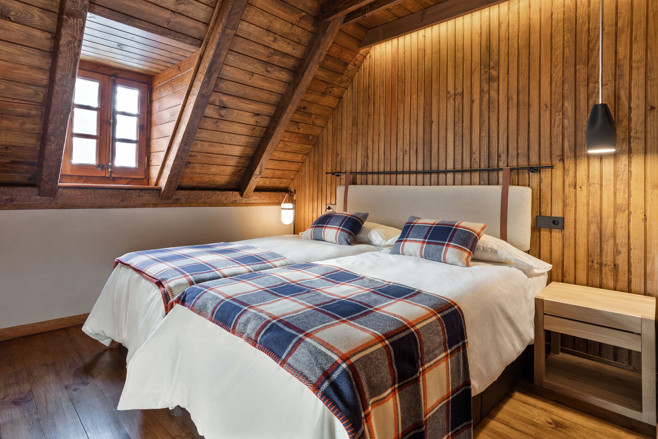 Habitación en un ático con paredes de madera, una cama doble con cabecera tapizada, mantas y almohadas de cuadros, lámparas de pared y ventana con moldura de madera en el techo.